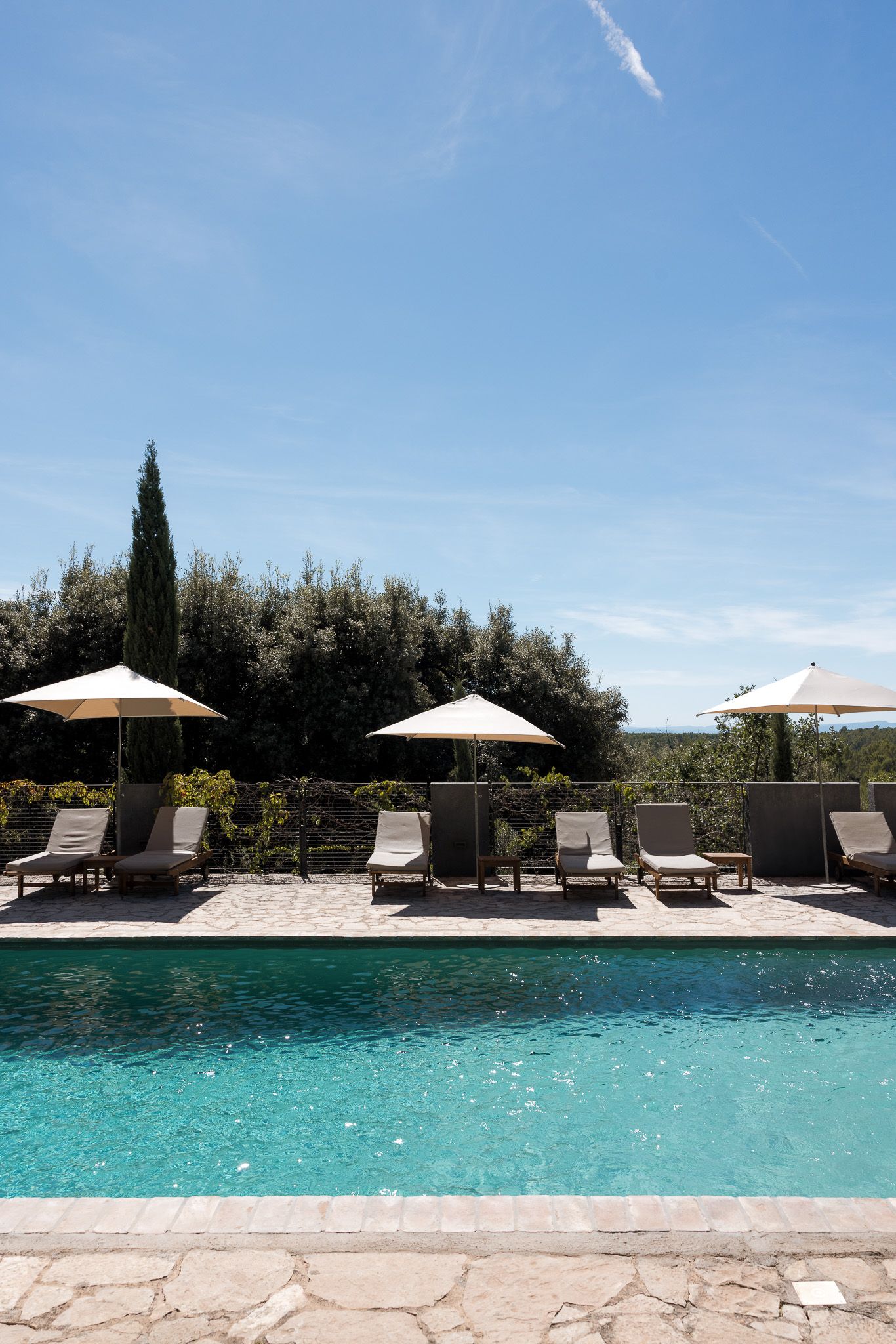 Luxury pool with lounge chairs and beige umbrellas surrounded by lush greenery under a clear blue sky.