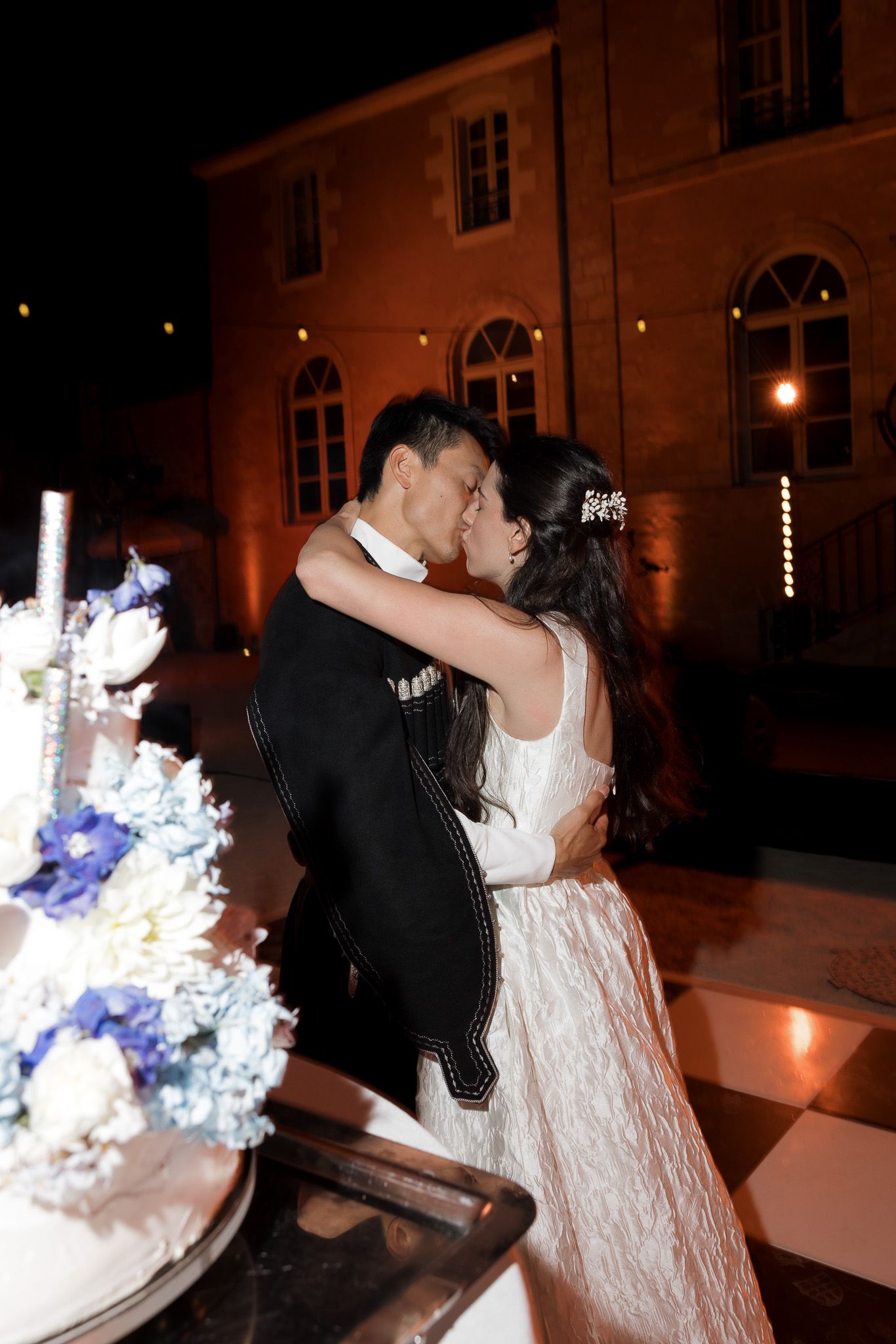 Bride and groom share a romantic kiss at night near a wedding cake with blue and white flowers at their outdoor reception.