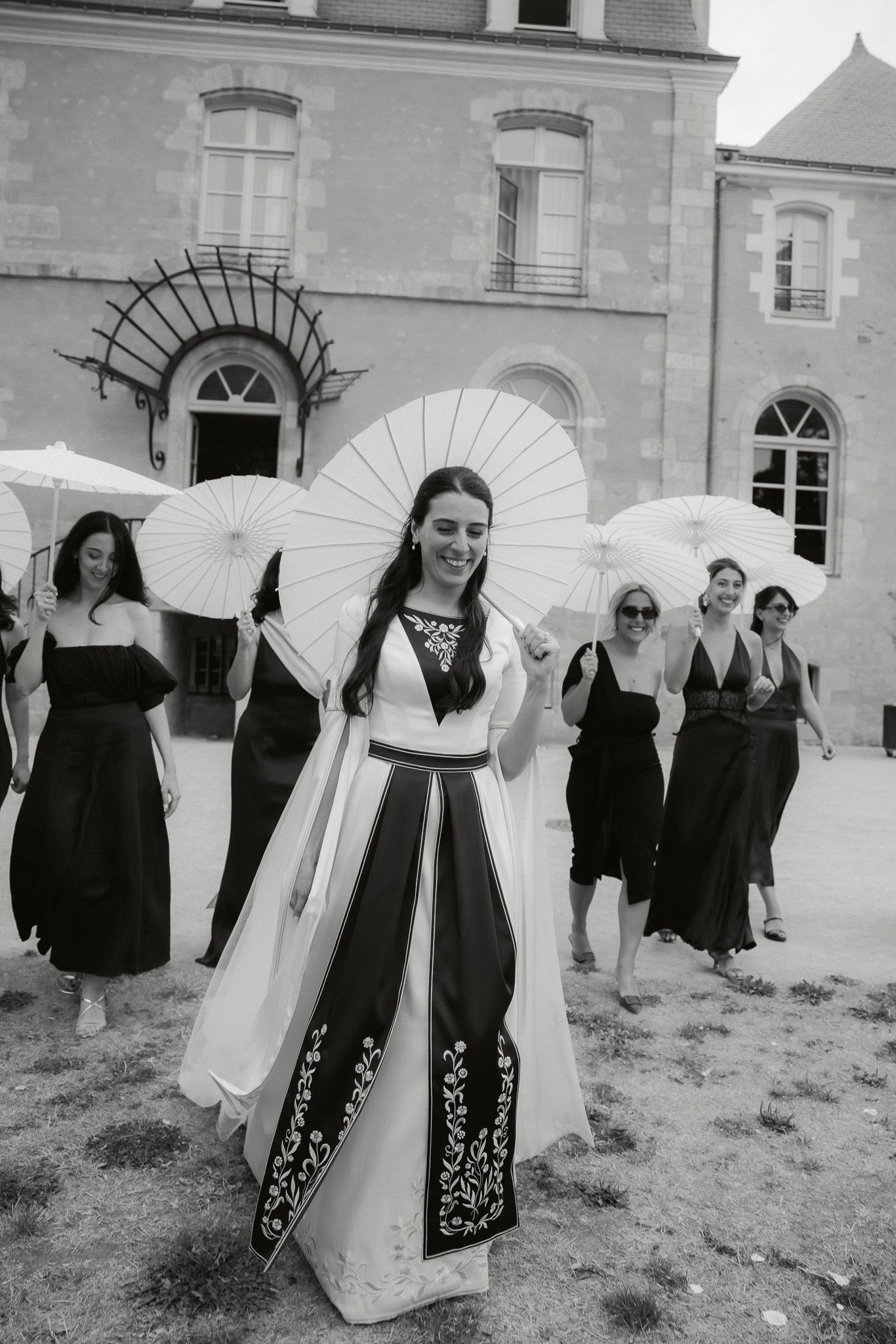 Bride in traditional embroidered gown holds parasol with bridesmaids in black dresses outside French château.