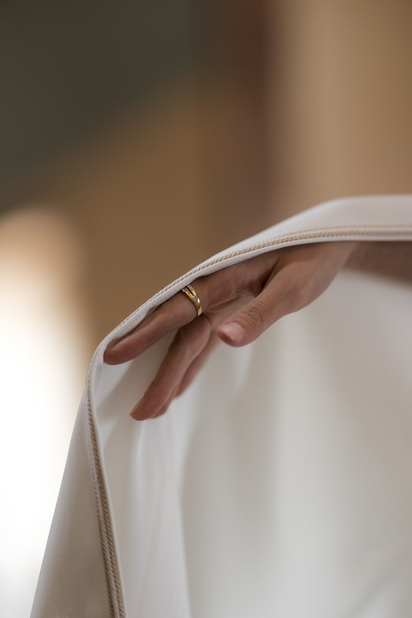 A hand with a gold ring delicately holds the edge of a white satin fabric in soft natural light.