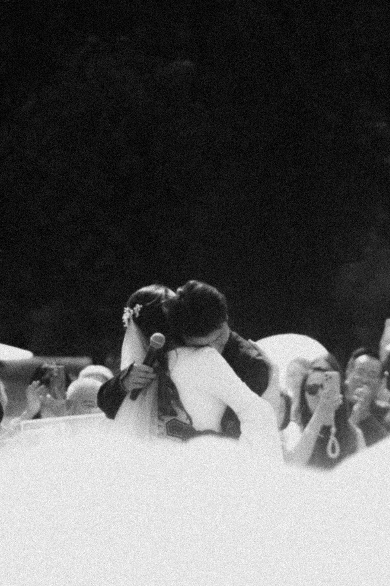 Black and white photo of a couple sharing a kiss at their wedding ceremony while guests celebrate around them.