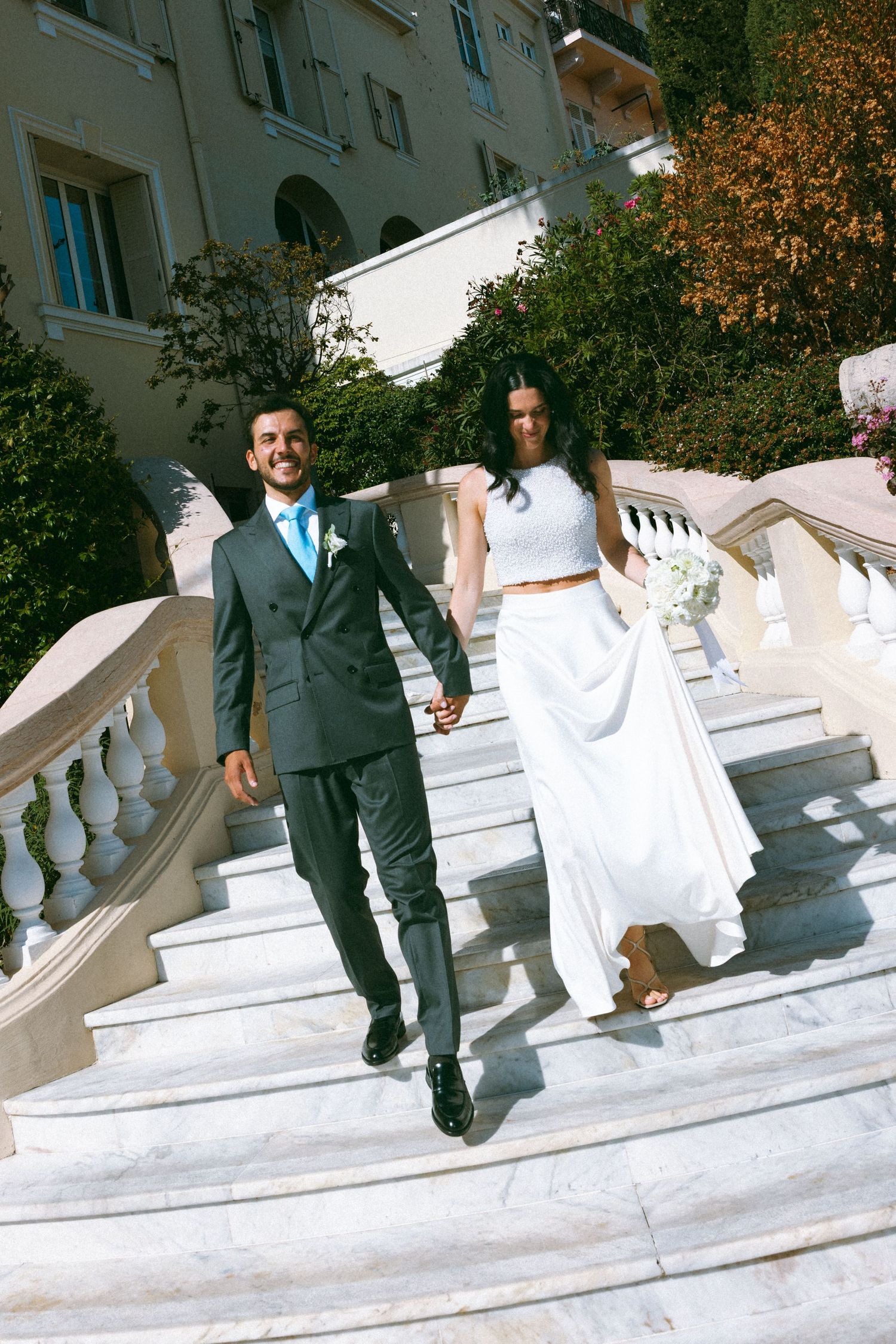 Bride and groom holding hands walking down elegant white marble stairs outside a grand building on their wedding day.