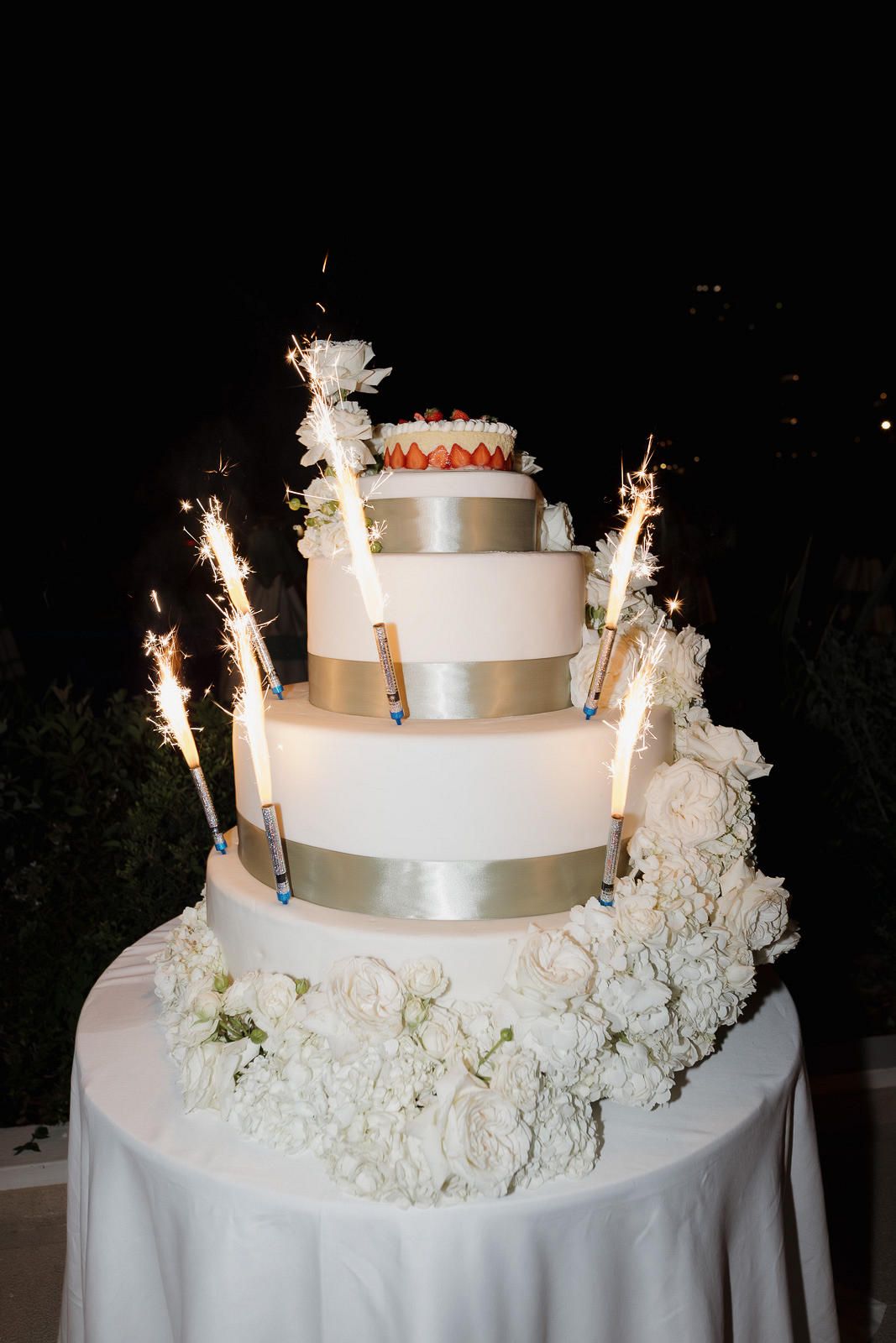 A white tiered wedding cake decorated with sparklers and white flowers sits on a table at night.