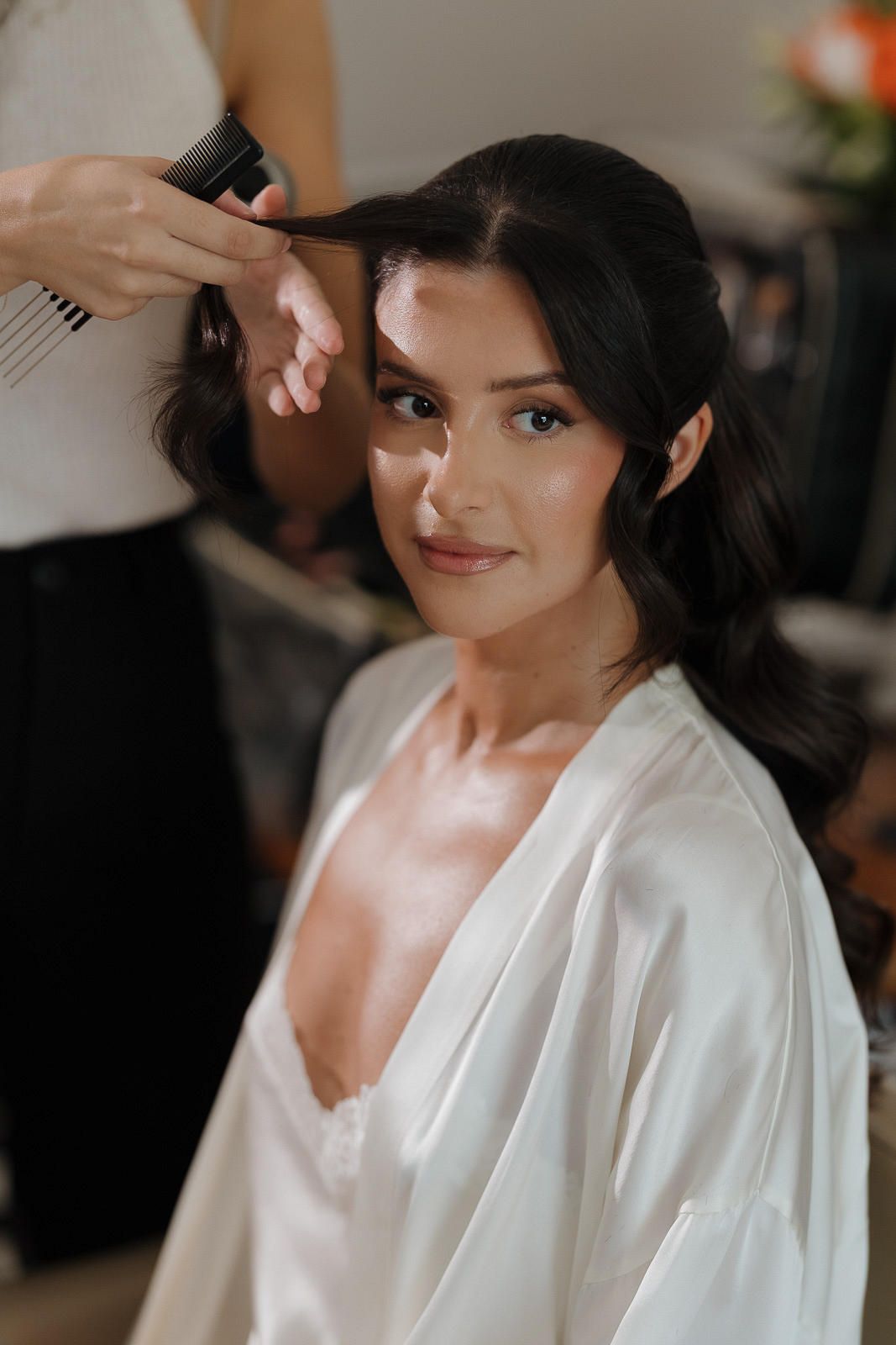 A bride getting ready for her wedding day wearing a white silk robe while having her hair and makeup done.