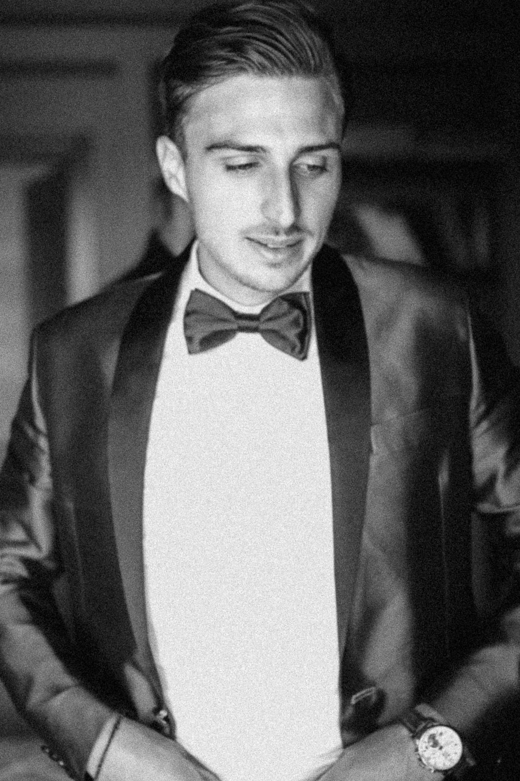 A black and white image shows a person in formal attire looking down at their wristwatch in dim lighting.