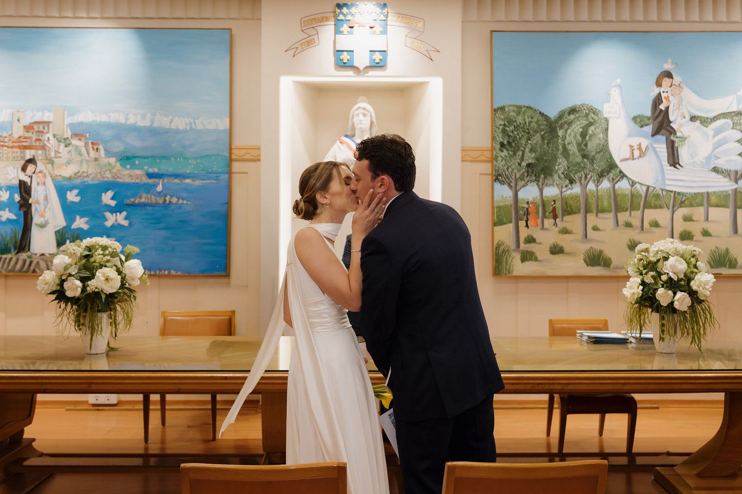 A wedding couple shares an intimate moment in front of beach-themed artwork displayed in a gallery setting.
