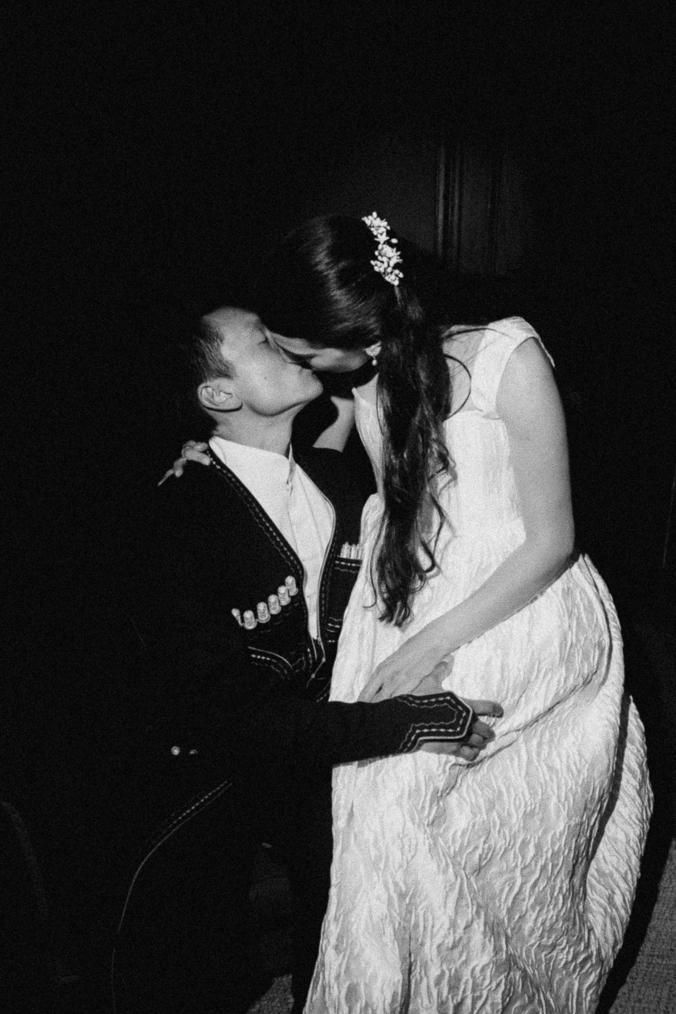 Black and white photo of a couple sharing a romantic kiss, bride in white dress with floral hair accessory.