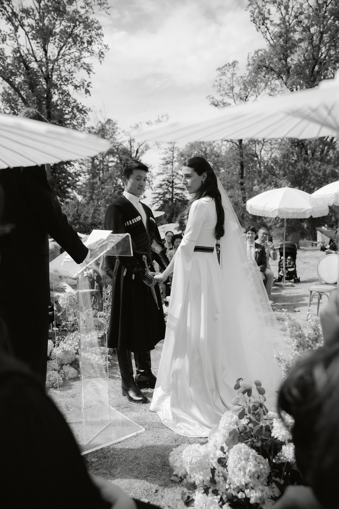 Black and white outdoor wedding ceremony with bride in flowing gown and groom exchanging vows among floral arrangements.