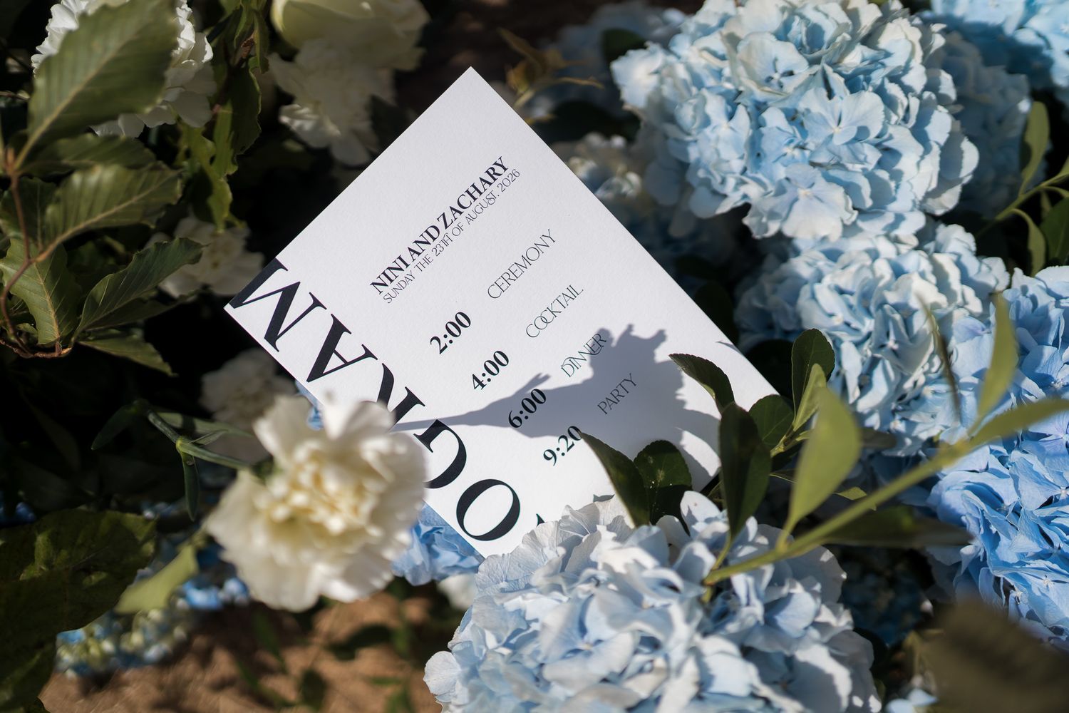 A white card with elegant typography rests among blue and white hydrangea flowers in a garden setting.