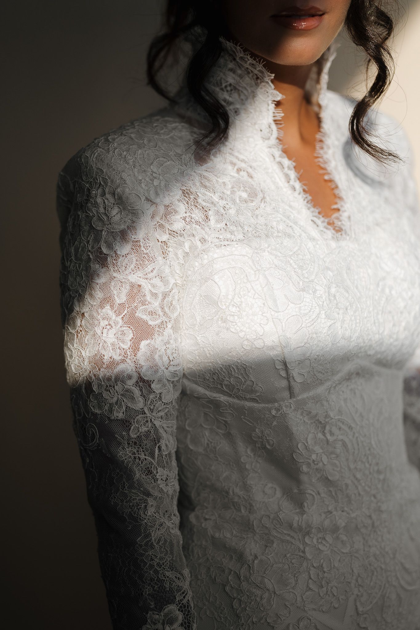 Bride wearing elegant white lace long-sleeve wedding dress with high neck, illuminated by soft natural light.