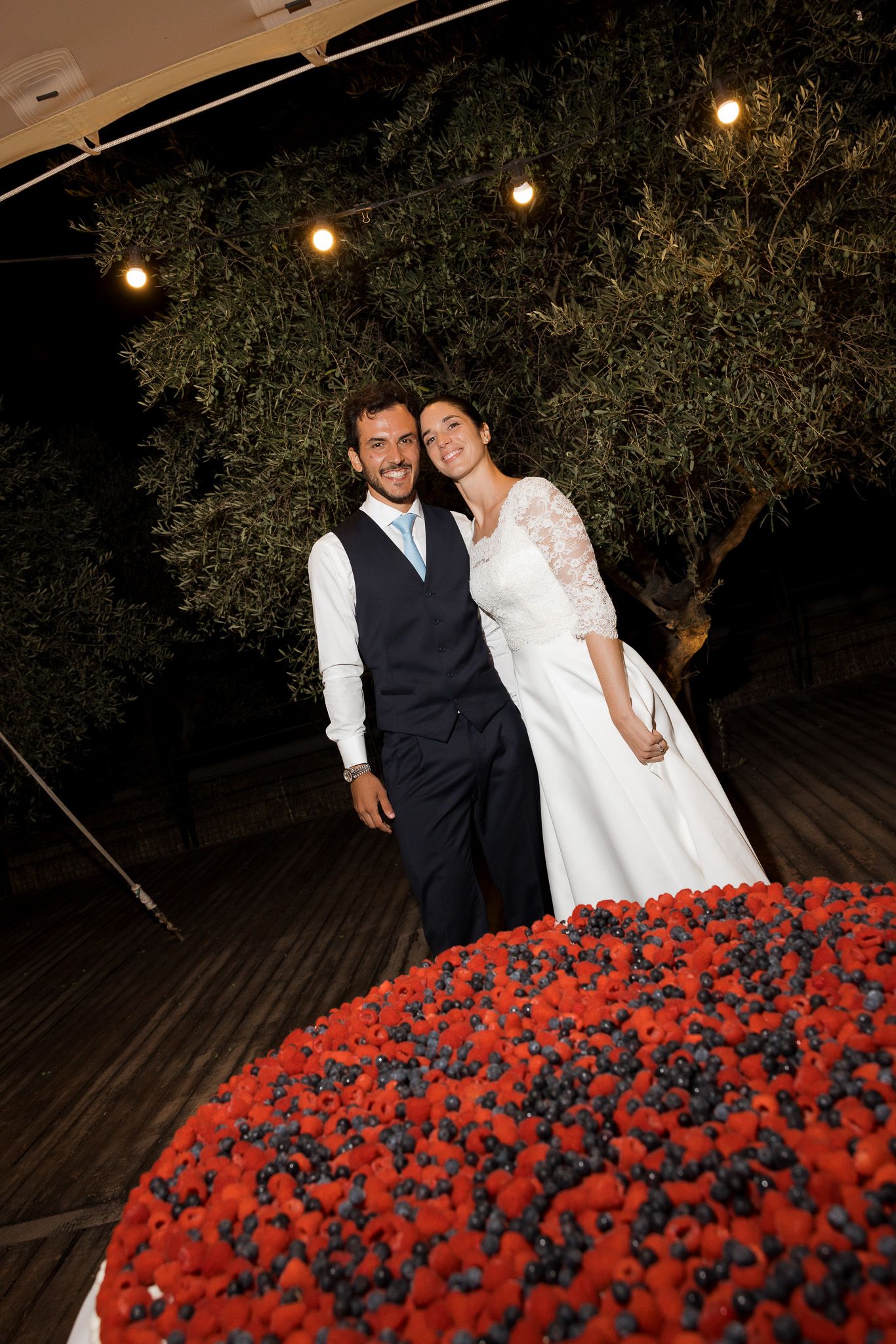 Bride and groom smiling together at night beside a heart-shaped berry cake decorated with strawberries and blueberries.