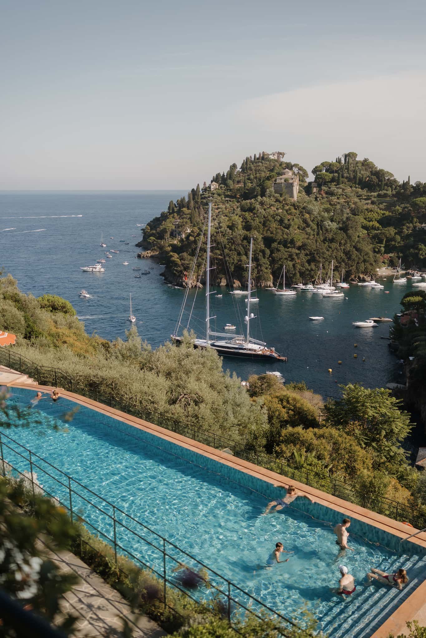 Luxury infinity pool overlooking a Mediterranean harbor filled with anchored boats and yachts on a sunny summer day.