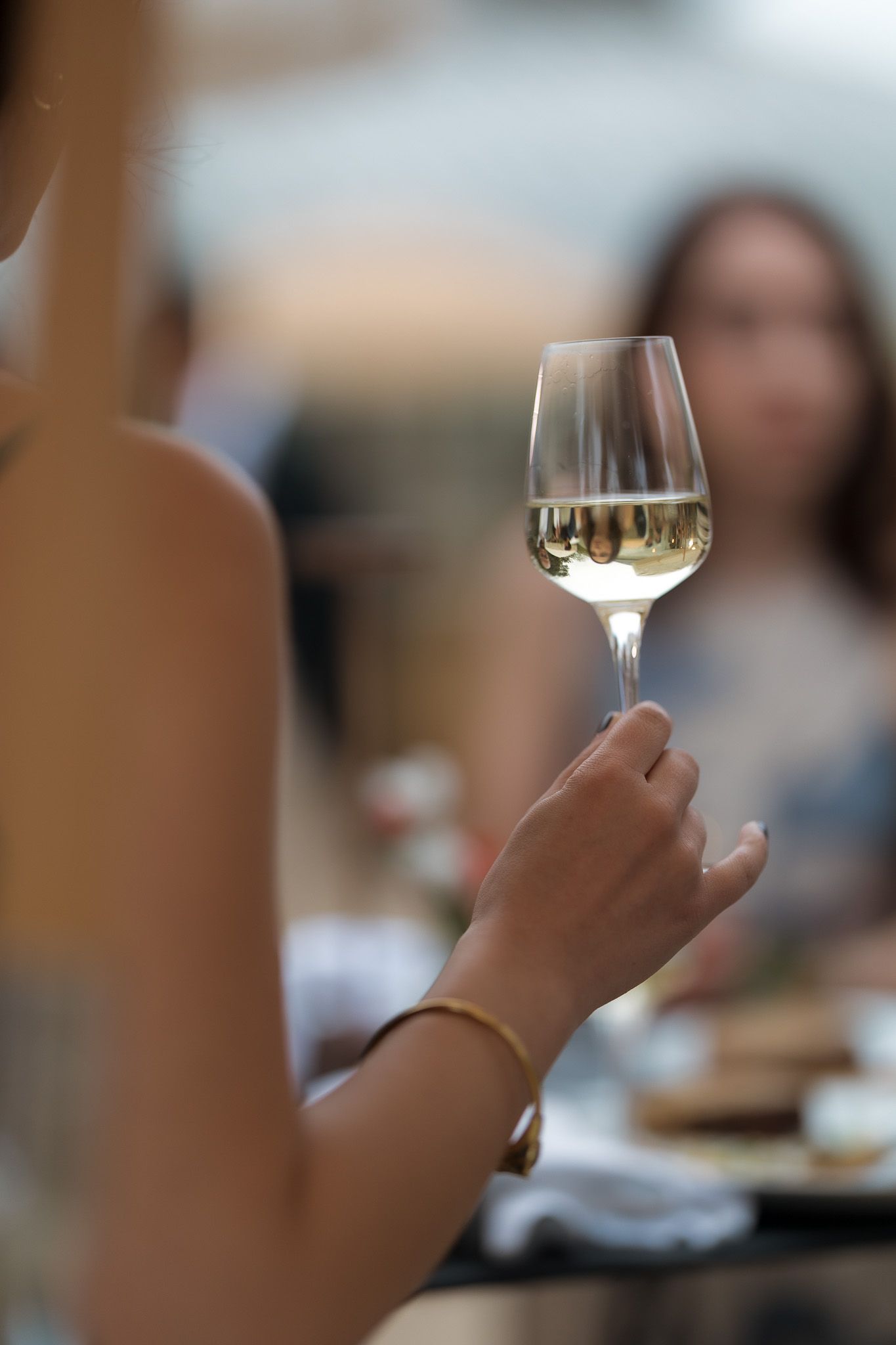 Woman holding a glass of white wine at an outdoor dining table with friends in the background.
