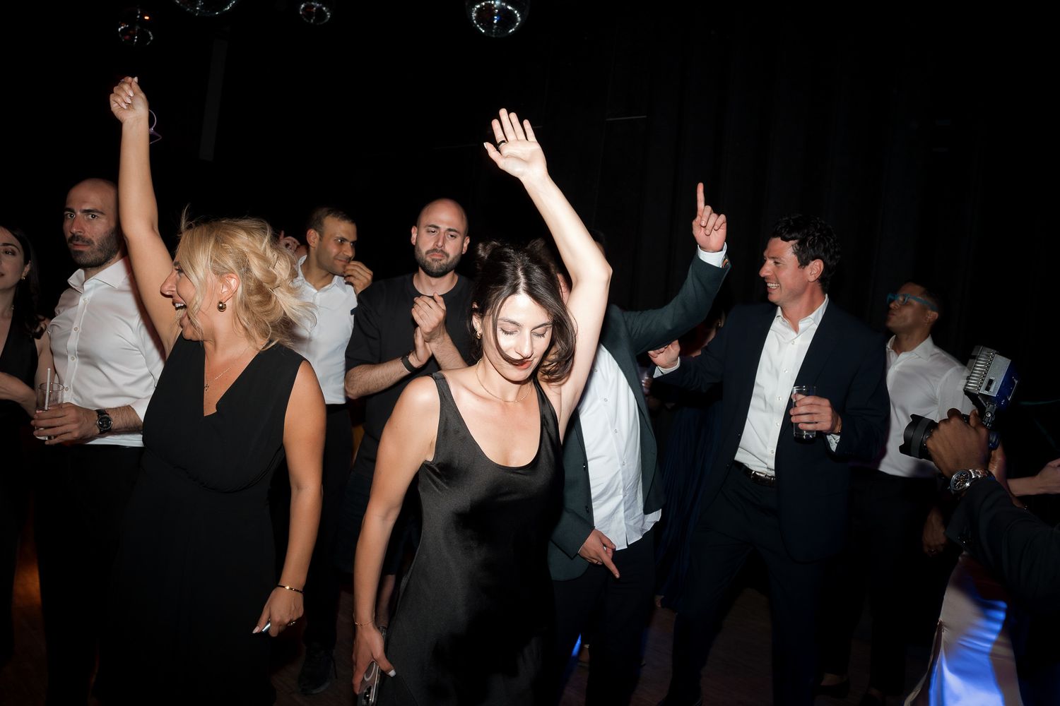 Guests dancing energetically at a wedding reception, wearing formal black and white attire in a dimly lit venue.