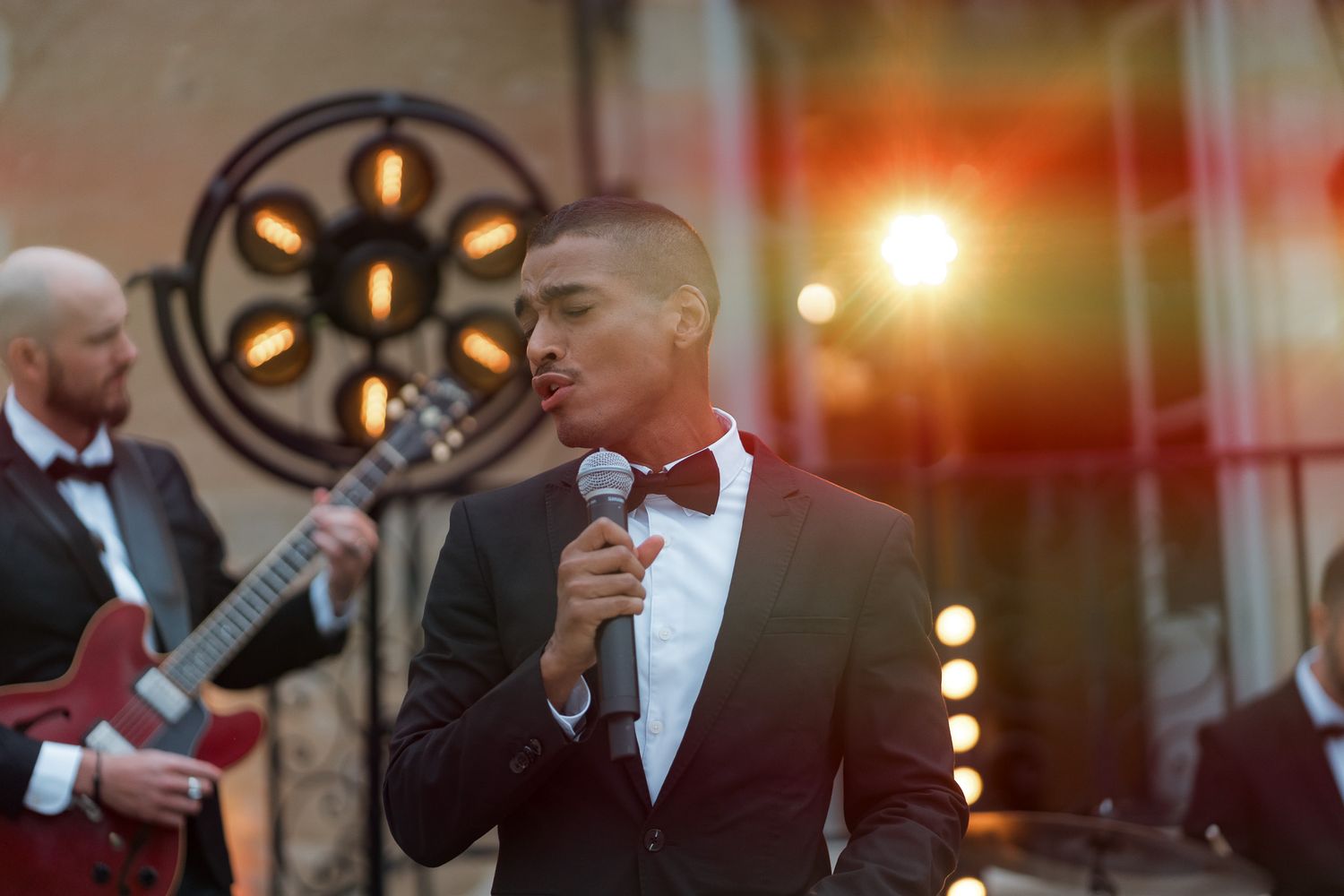 Young male singer in tuxedo performing on stage with microphone, guitarist in background at outdoor event.