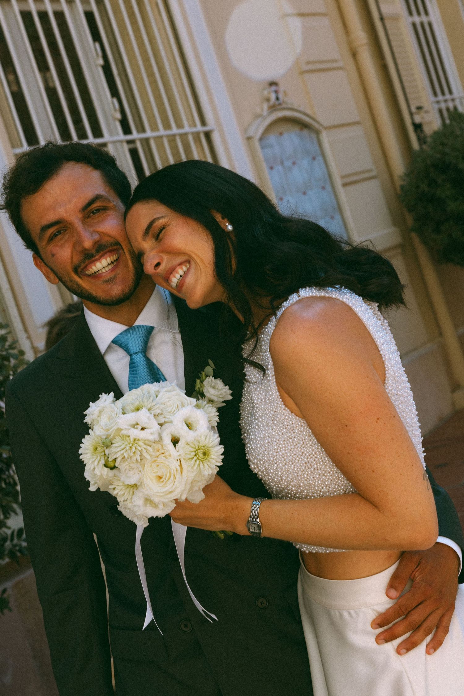 Happy newlyweds embrace and smile, bride holding white flower bouquet in elegant wedding attire outdoors.