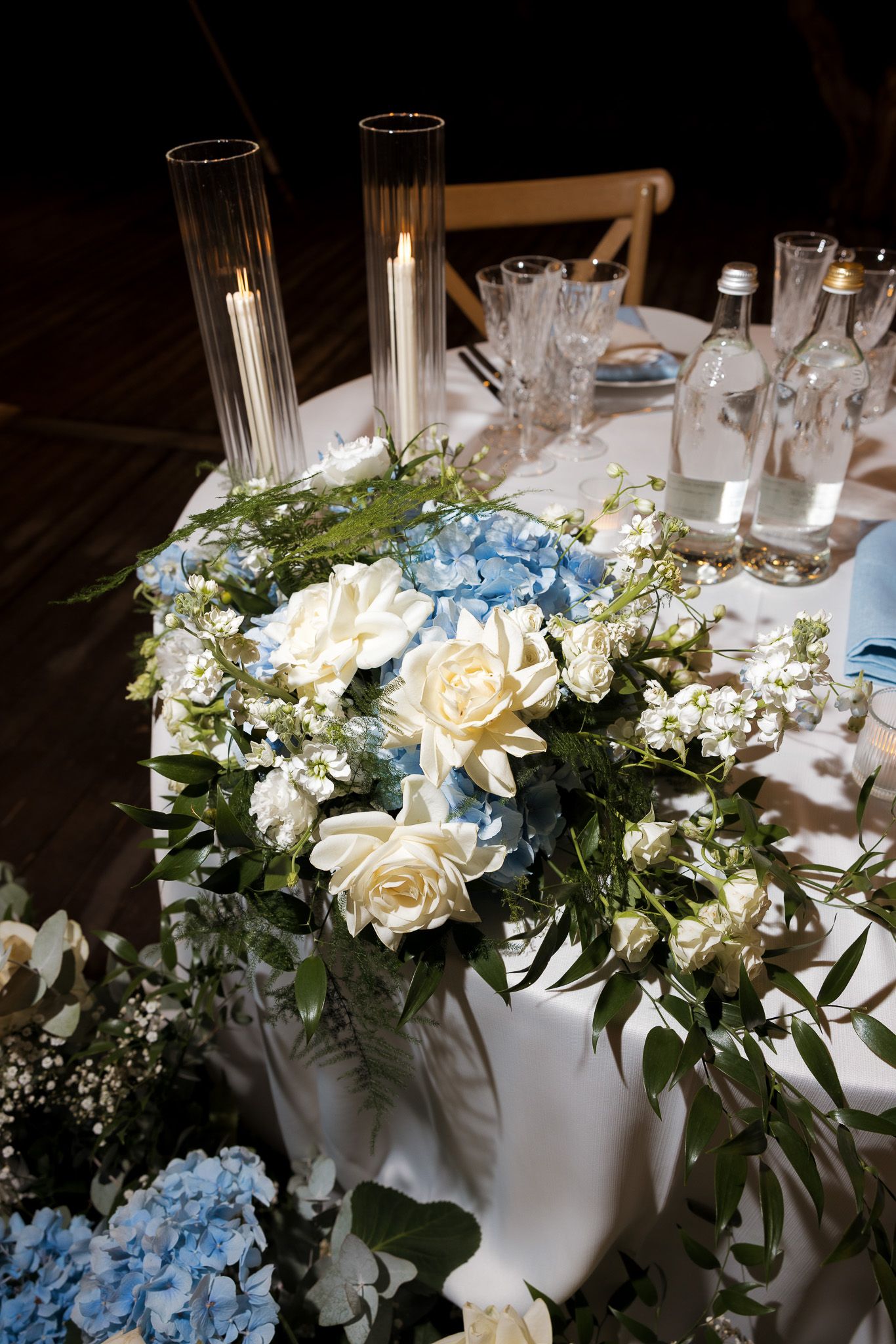 Elegant wedding table centerpiece with white roses, blue hydrangeas, greenery, and tall candles on a white tablecloth.