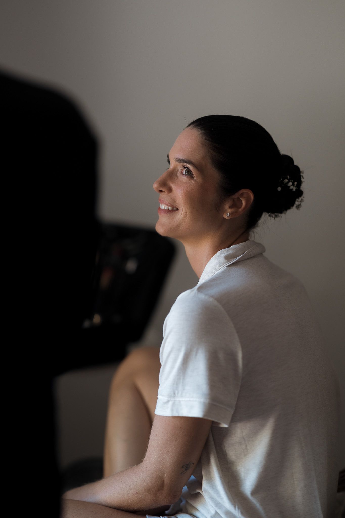 Woman in white polo shirt with hair in bun smiling, seated in softly lit room.
