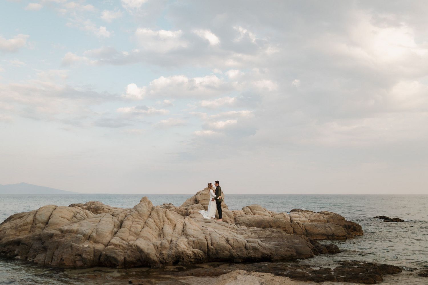 Two silhouettes stand on a rocky coastal outcrop overlooking the ocean during a cloudy day.