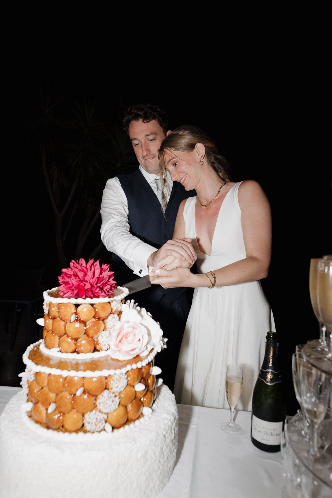 A wedding couple cutting their three-tiered cream and gold croquembouche cake decorated with pink flowers.