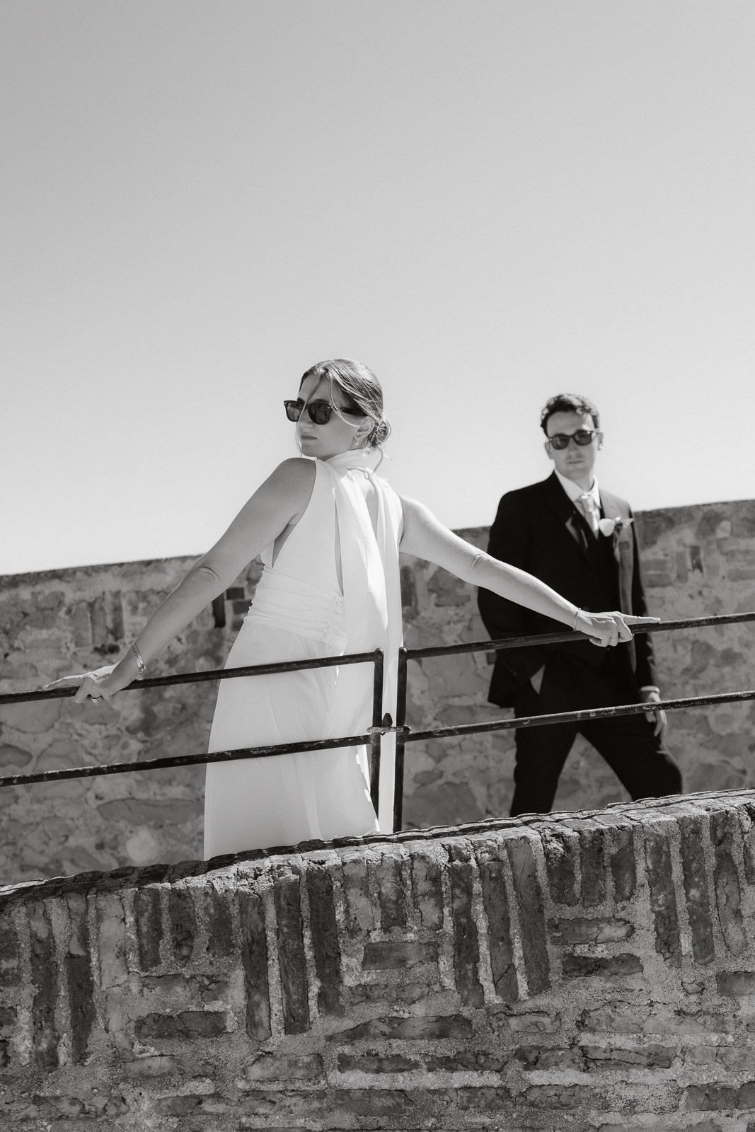 A black and white image shows two people walking along an old stone wall in formal attire.