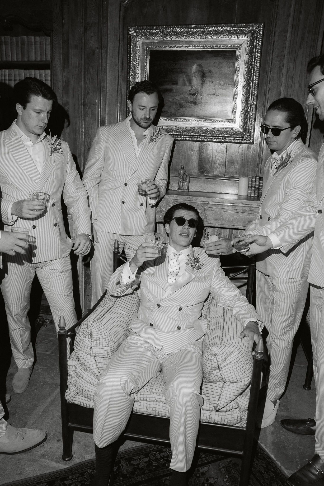 Groom in white double-breasted suit and sunglasses seated in chair surrounded by groomsmen in matching suits at wedding.
