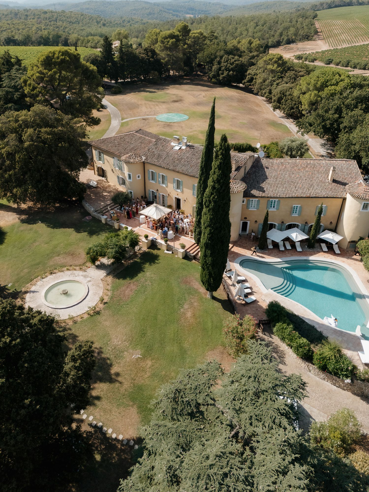 Aerial view of a stunning Provençal villa with pool, fountain, and lush gardens surrounded by vineyards.