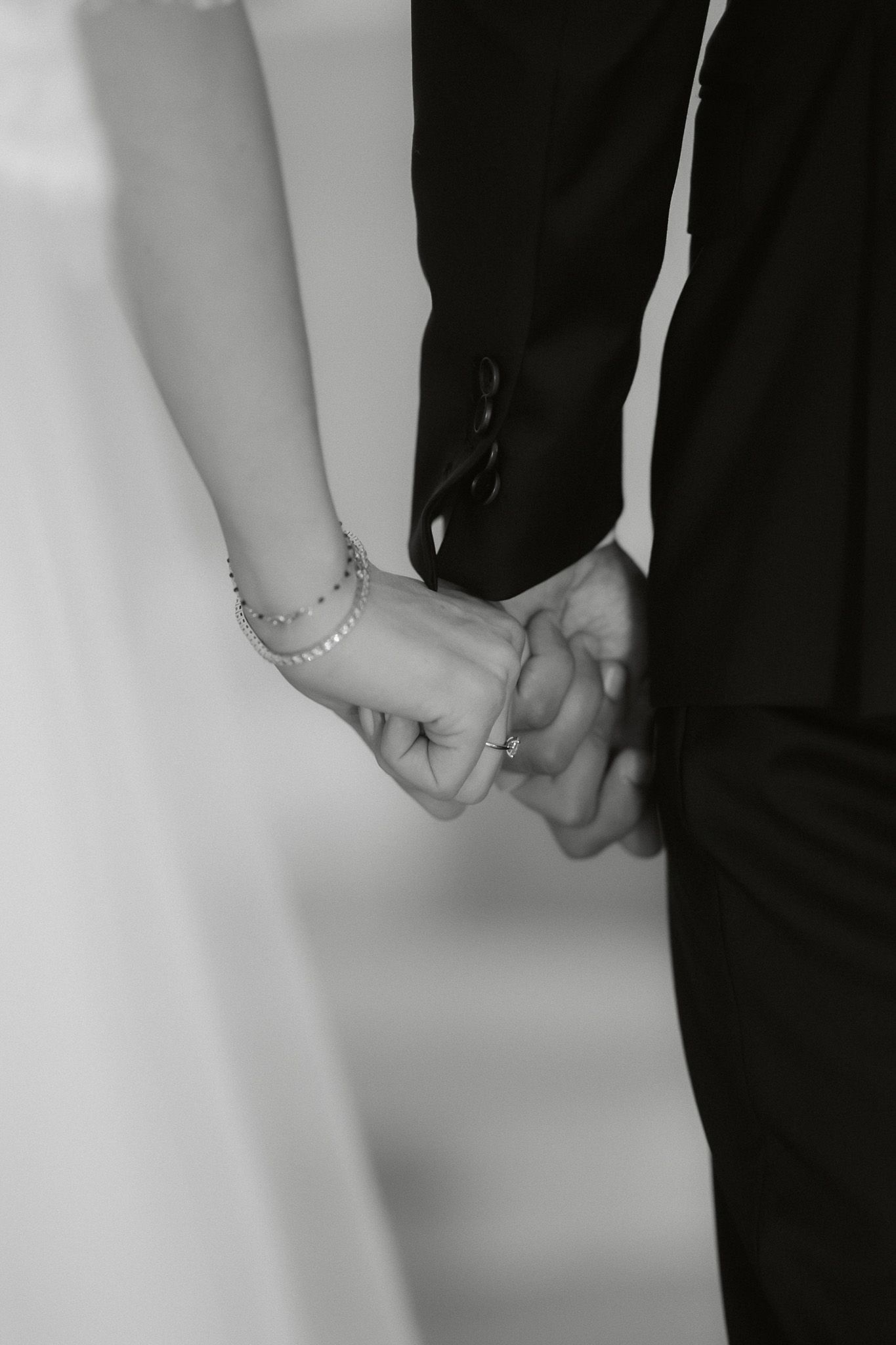 Black and white photo of a bride and groom holding hands, showing wedding rings and a delicate bracelet.