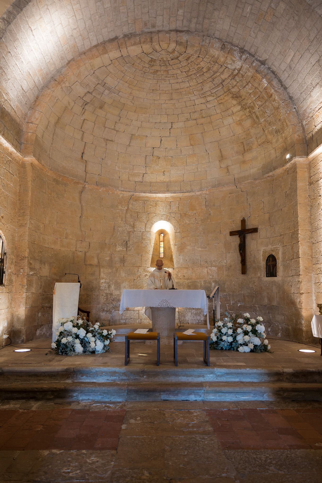 Ancient Romanesque church interior with stone barrel vault, altar, white flowers, and wooden cross.