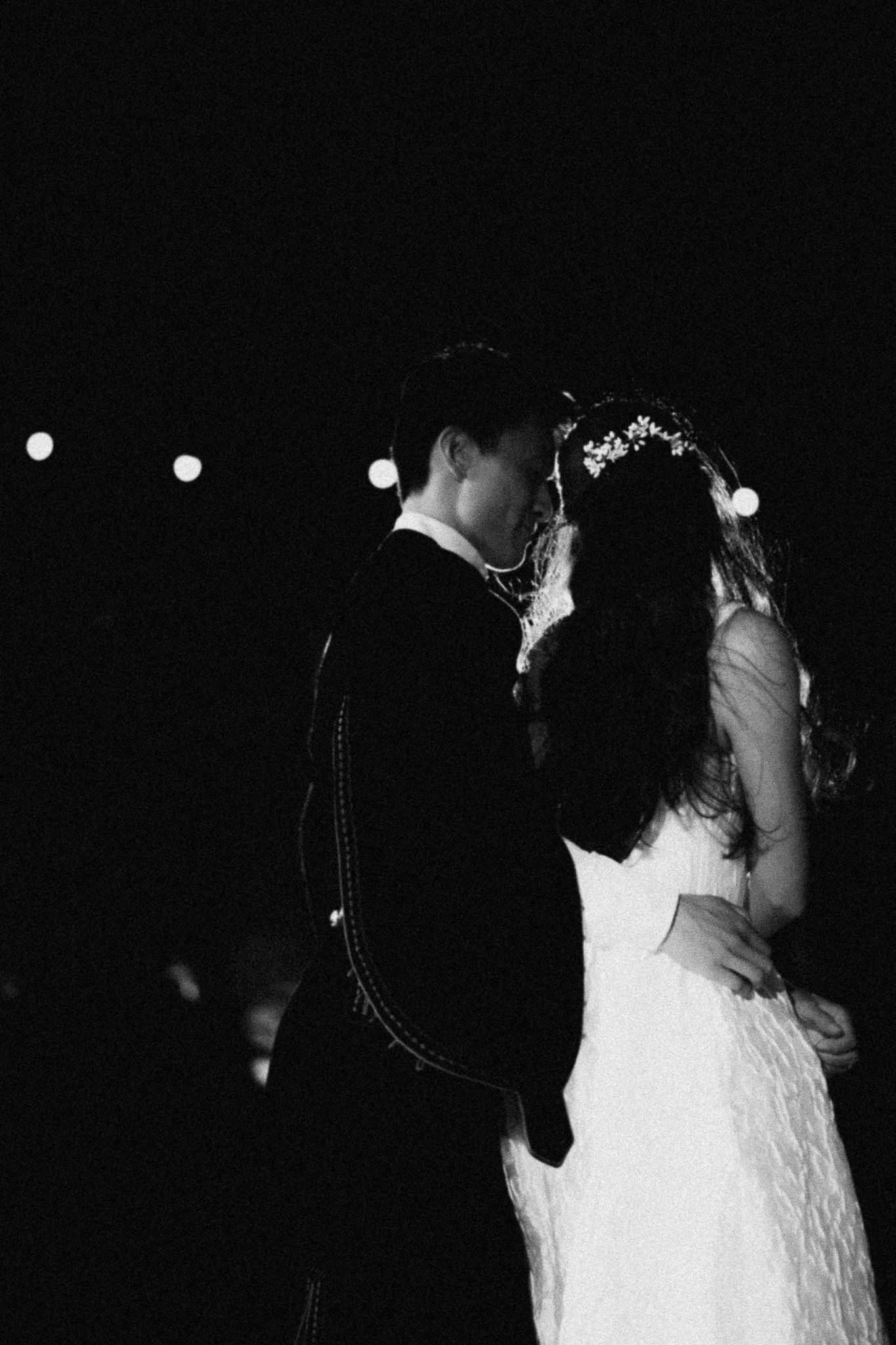 Black and white photo of a bride and groom sharing their first dance under string lights at a romantic wedding reception.