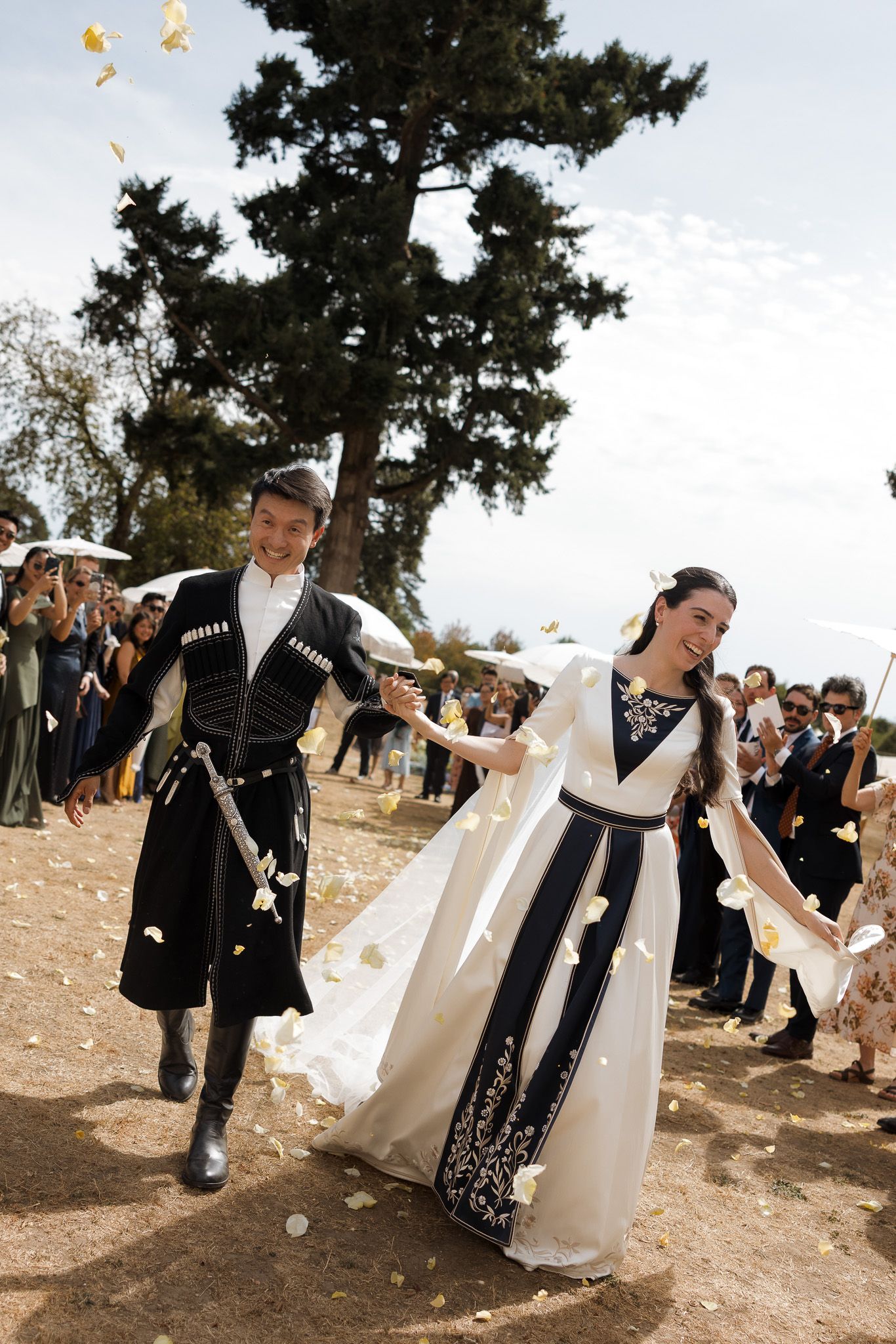 Couple in traditional Georgian wedding attire walking joyfully as guests toss flower petals outdoors.