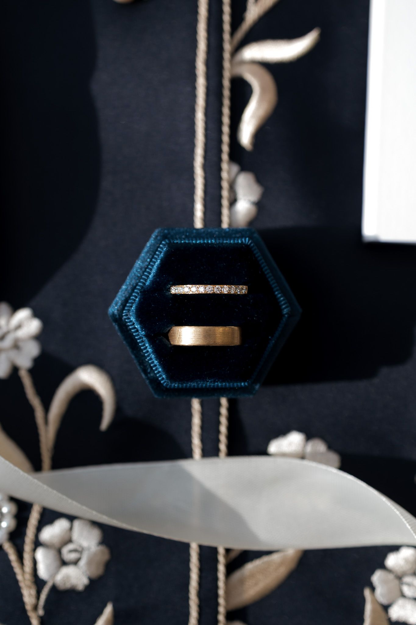 Two wedding rings displayed in a teal hexagonal velvet ring box against dark floral embroidered background.
