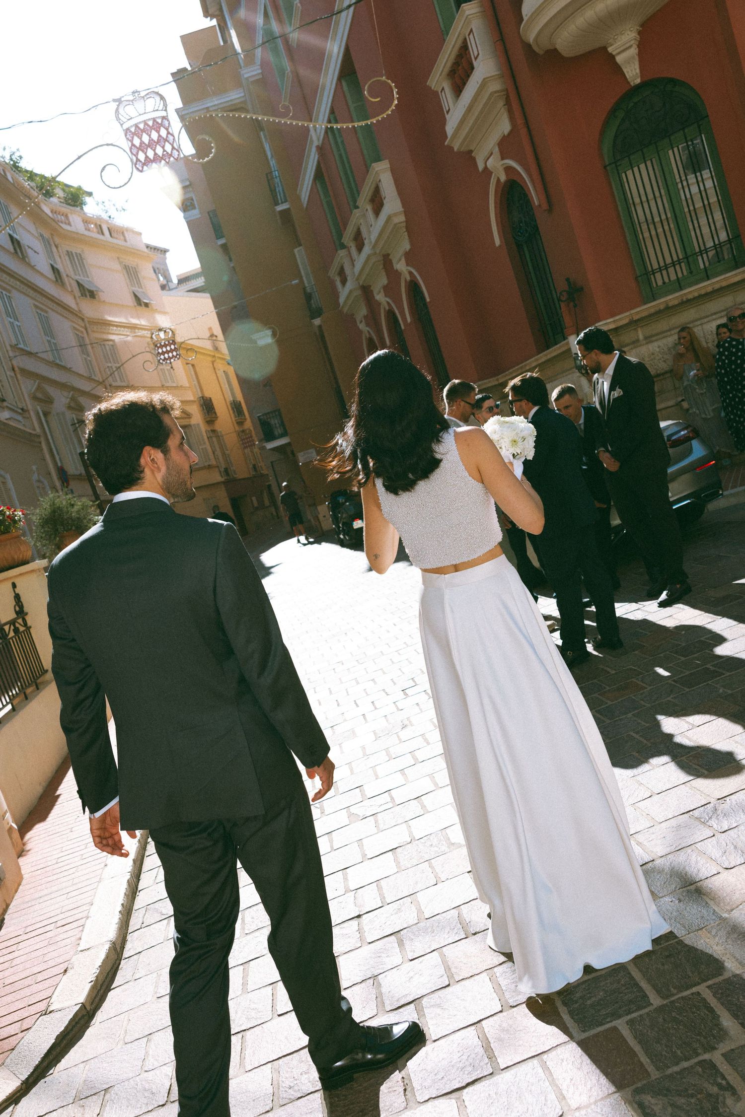 Bride in white gown and groom in black suit walk toward wedding guests on a sunlit cobblestone street in Italy.