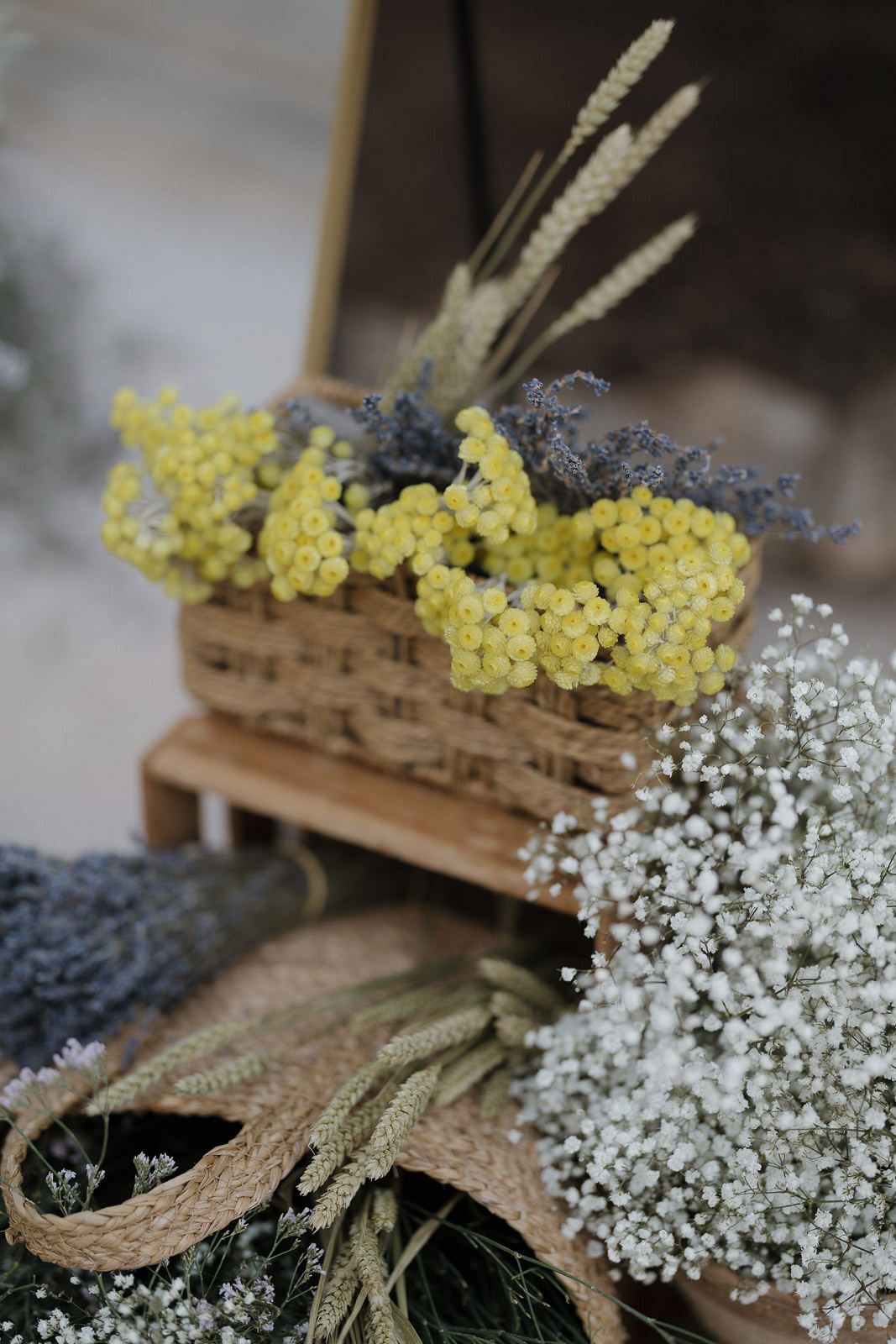 Rustic woven basket arrangement with yellow yarrow flowers, white baby's breath, and dried wheat stems on wooden surface.