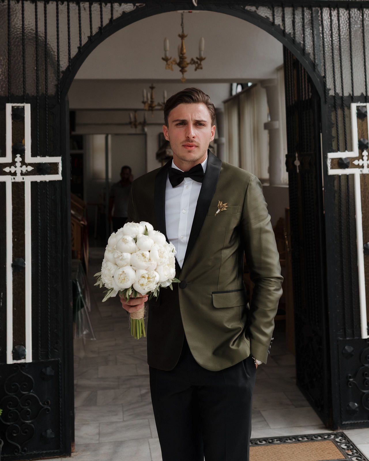 A person in a dark bow tie and olive green suit jacket holds a white rose bouquet while standing in an elegant doorway.