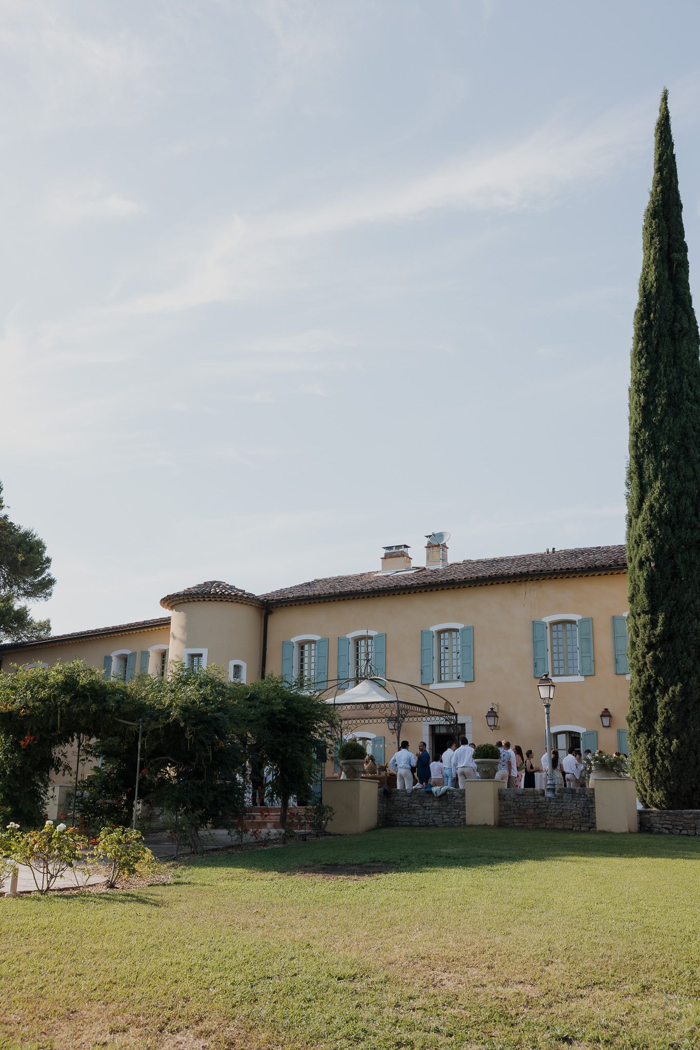 Elegant French villa with cypress tree, blue shutters, and guests gathered on the terrace during a sunny outdoor event.