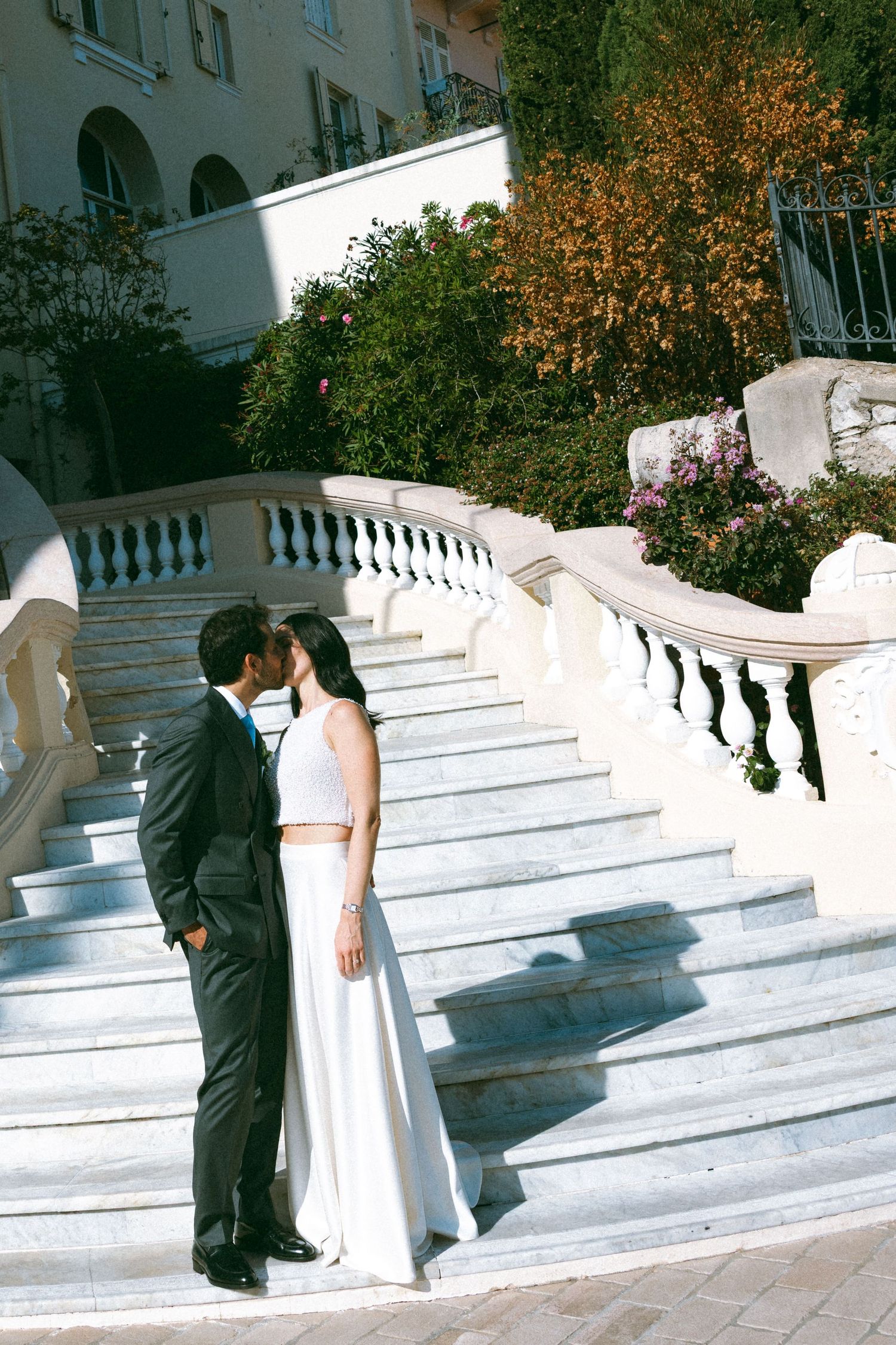 A bride and groom share a kiss on elegant white marble stairs surrounded by lush greenery at an outdoor wedding venue.