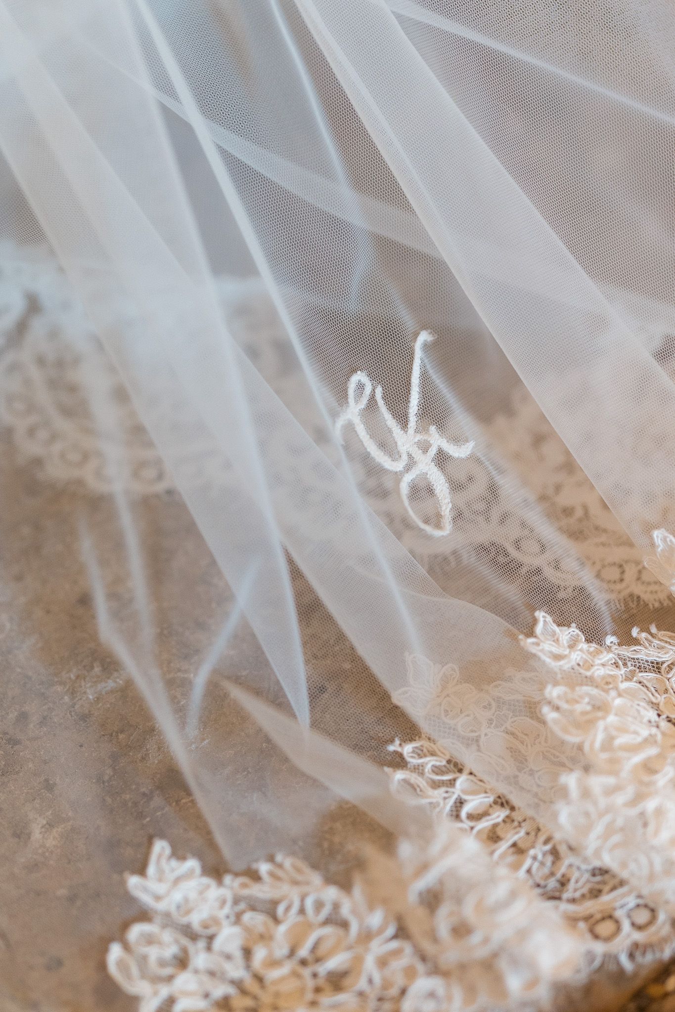 Close-up of a delicate white bridal veil with lace trim and embroidered monogram initials on a marble surface.
