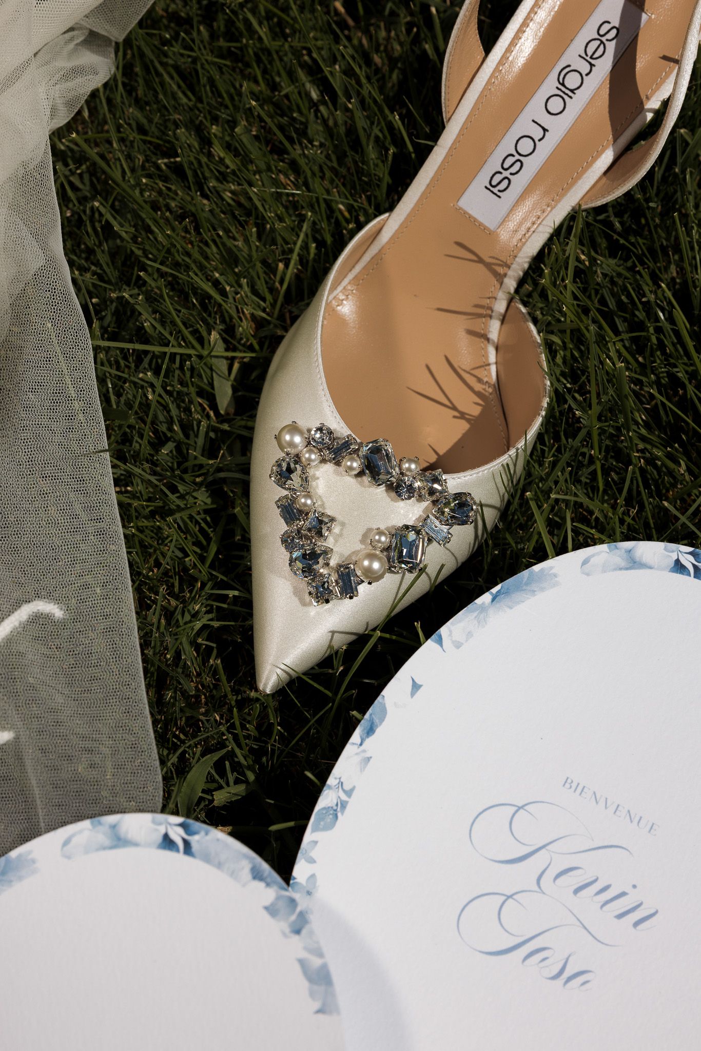 Sergio Rossi bridal heels with jeweled brooch beside Kevin and Jose wedding programs on green grass.