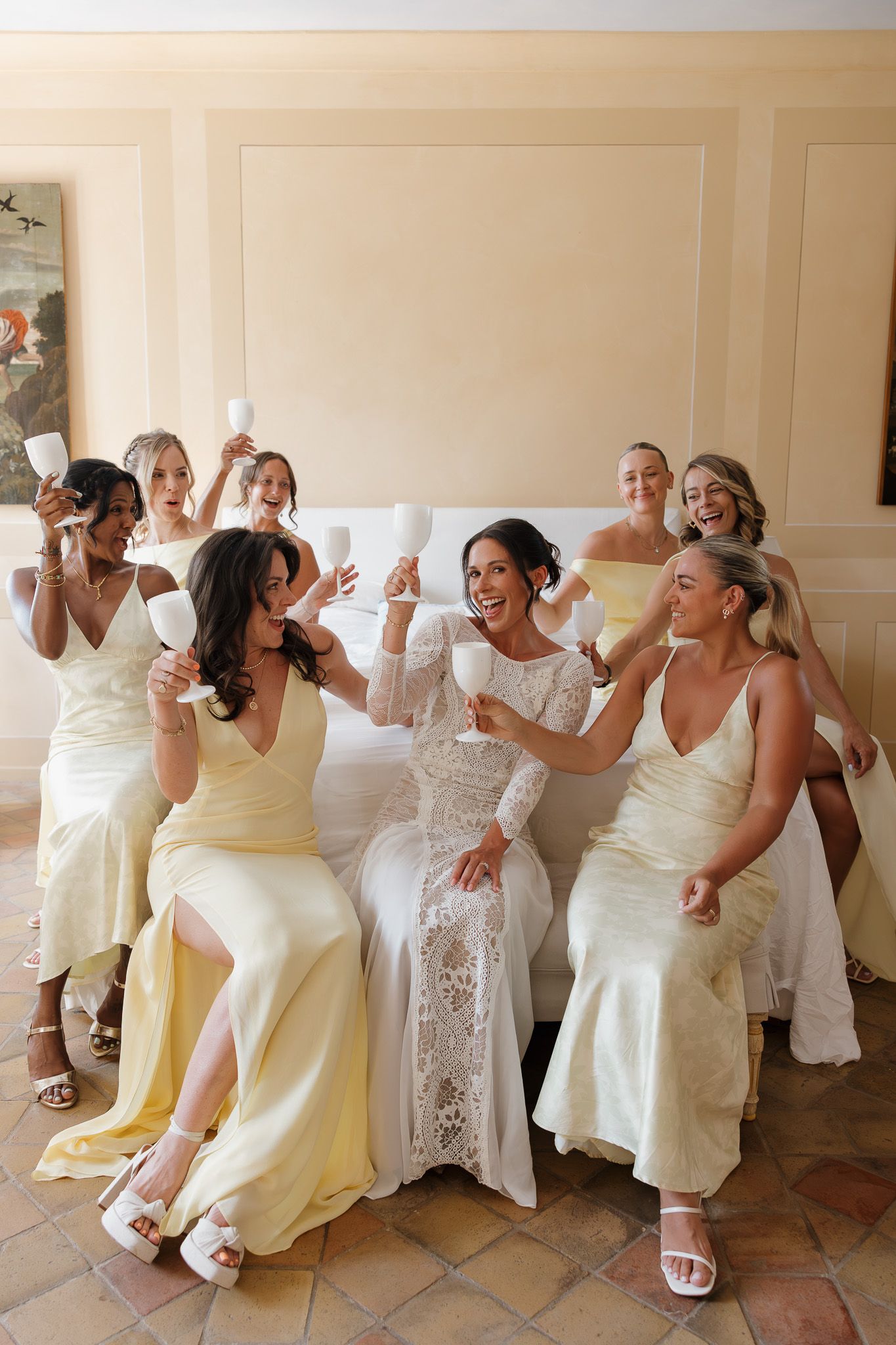 Bride in white lace gown celebrates with bridesmaids in yellow dresses, raising champagne glasses together.