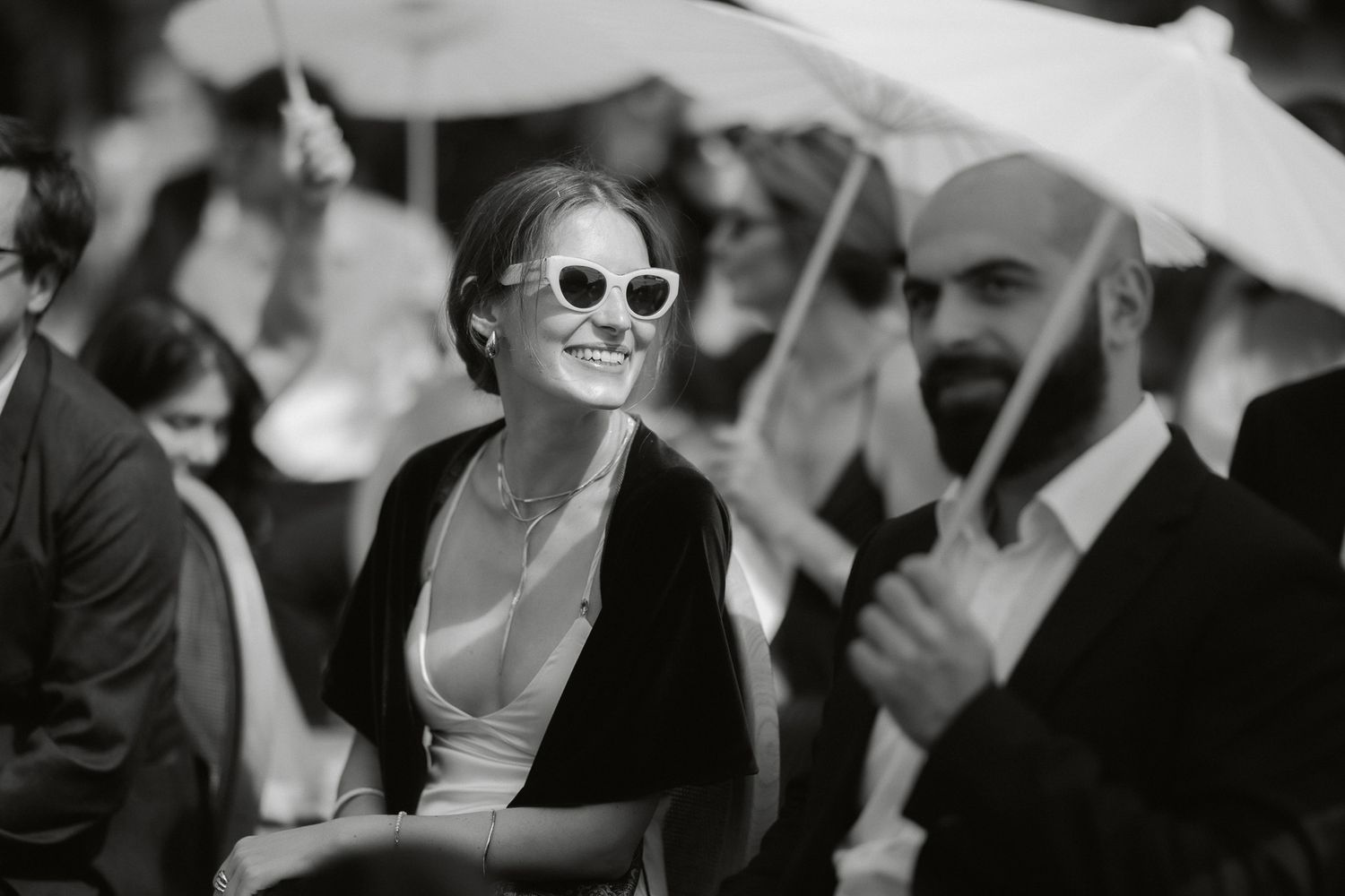 Black and white photo of a smiling woman in cat-eye sunglasses at an outdoor wedding ceremony.