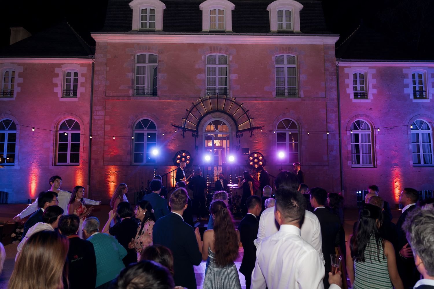 Outdoor evening wedding reception with guests dancing in front of an illuminated historic building with colorful stage lights.