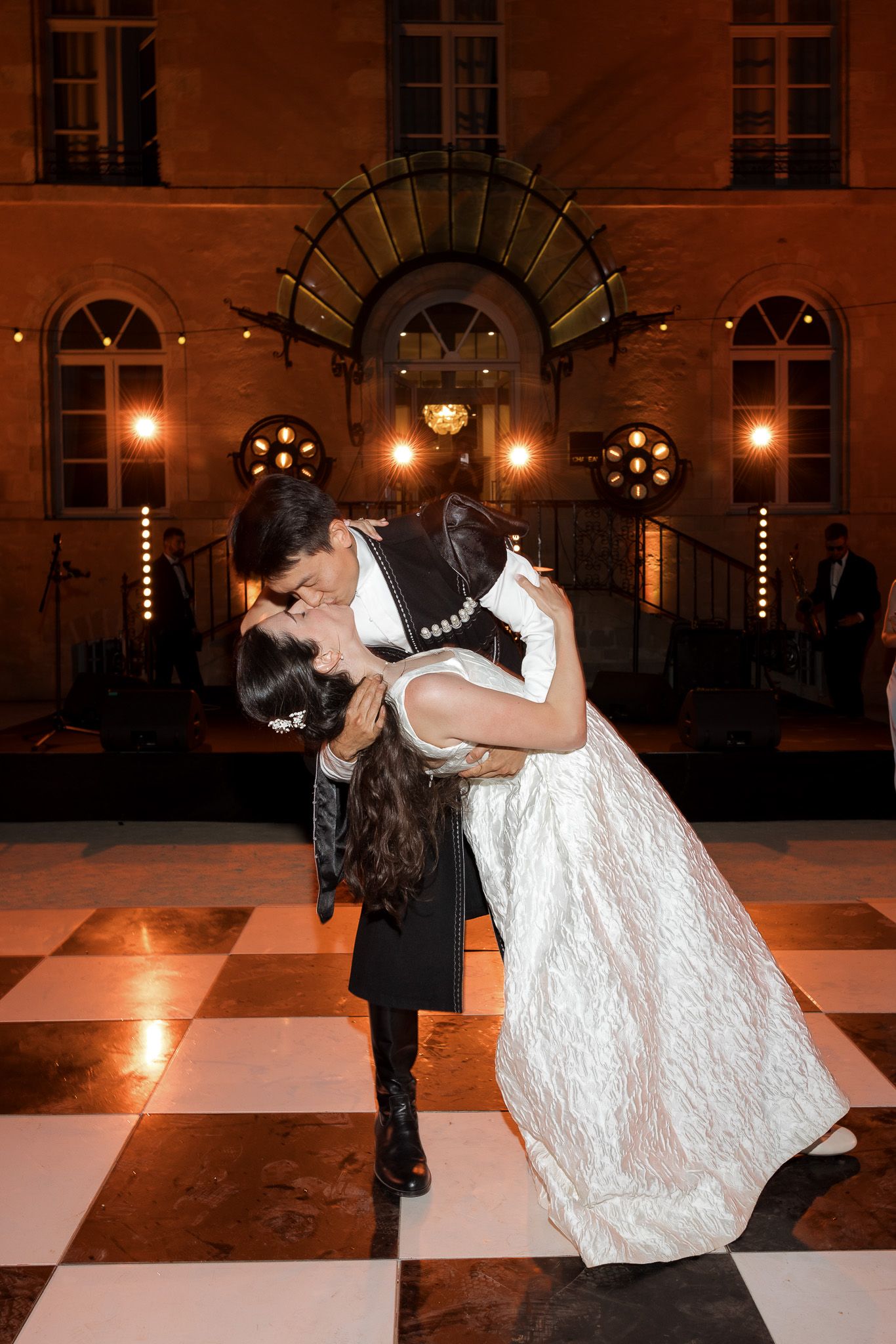 Couple sharing a romantic dip kiss on a black and white checkered dance floor outside an elegant lit building at night.