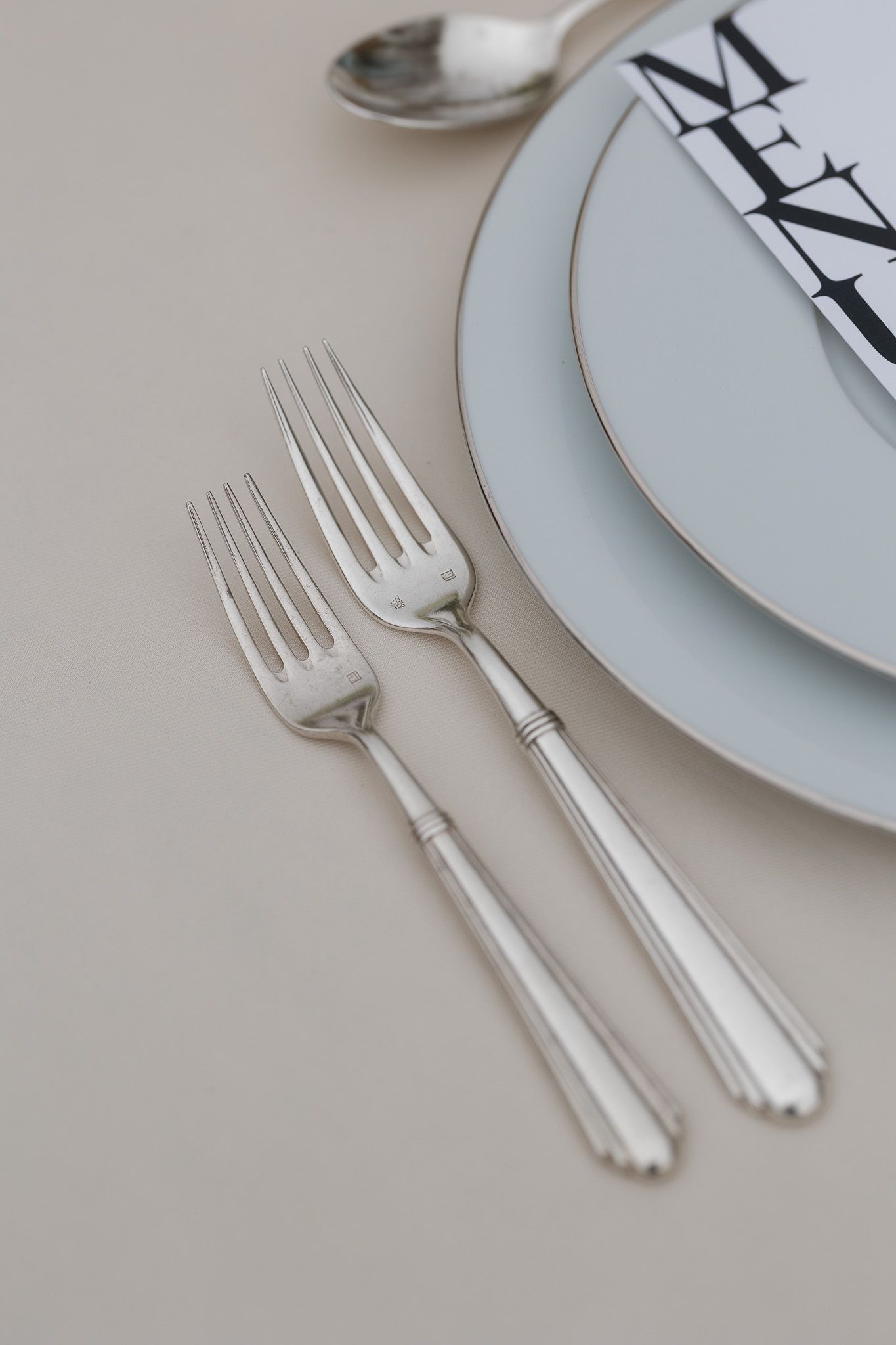 Elegant silver forks beside stacked white plates with gold rim and menu card on beige tablecloth.
