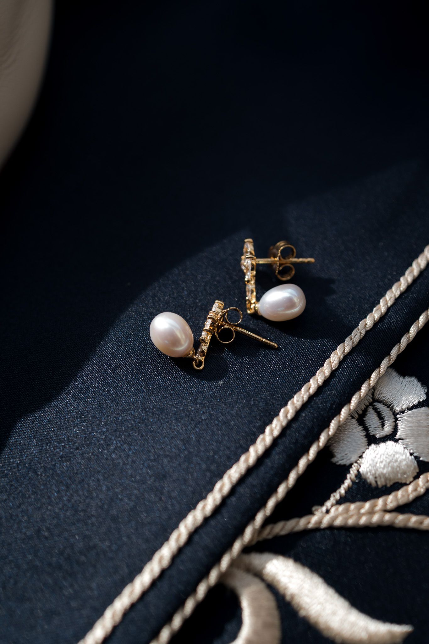 Gold bar stud earrings with crystals and white freshwater pearl drops on dark navy fabric.
