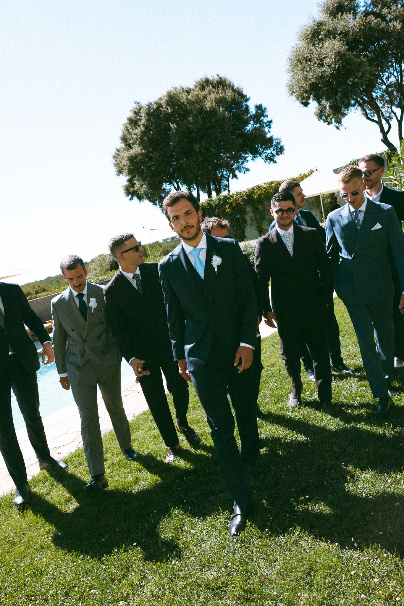 Groom leads groomsmen in suits across green lawn near pool on sunny wedding day outdoors.
