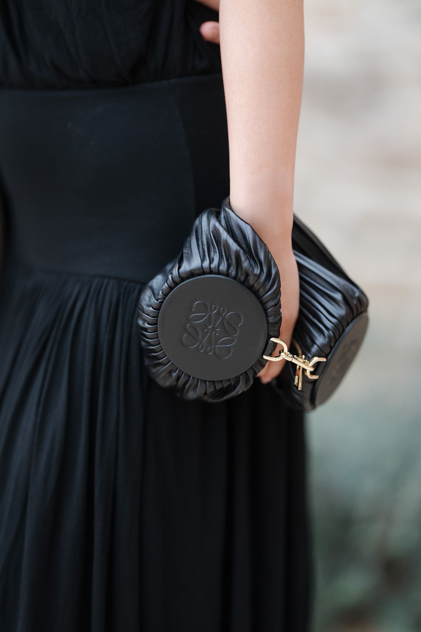 Woman holding a black Loewe ruched leather clutch bag with gold hardware against a pleated black dress.