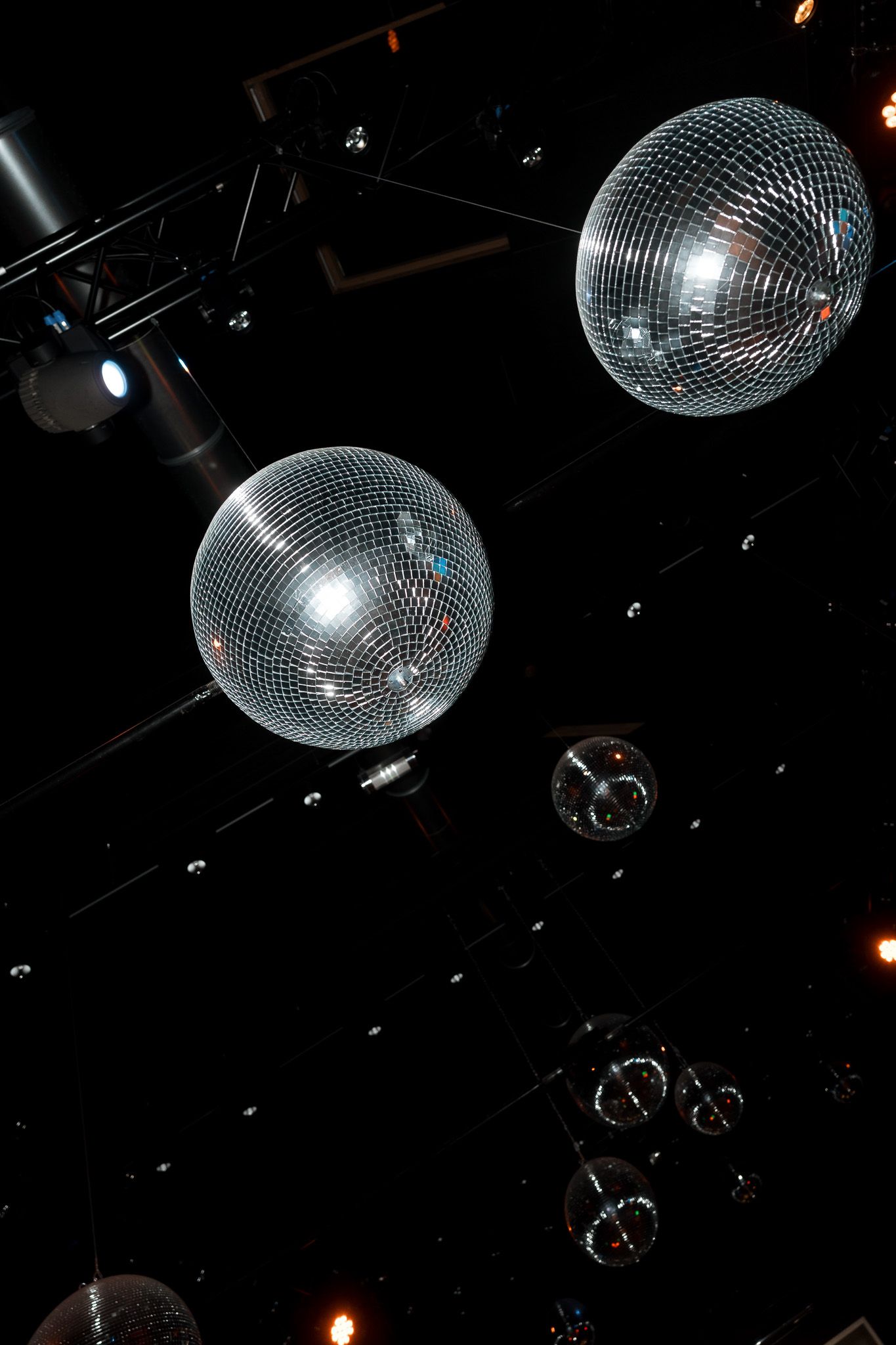 Multiple shiny disco mirror balls hanging from a dark nightclub ceiling with stage lighting equipment.
