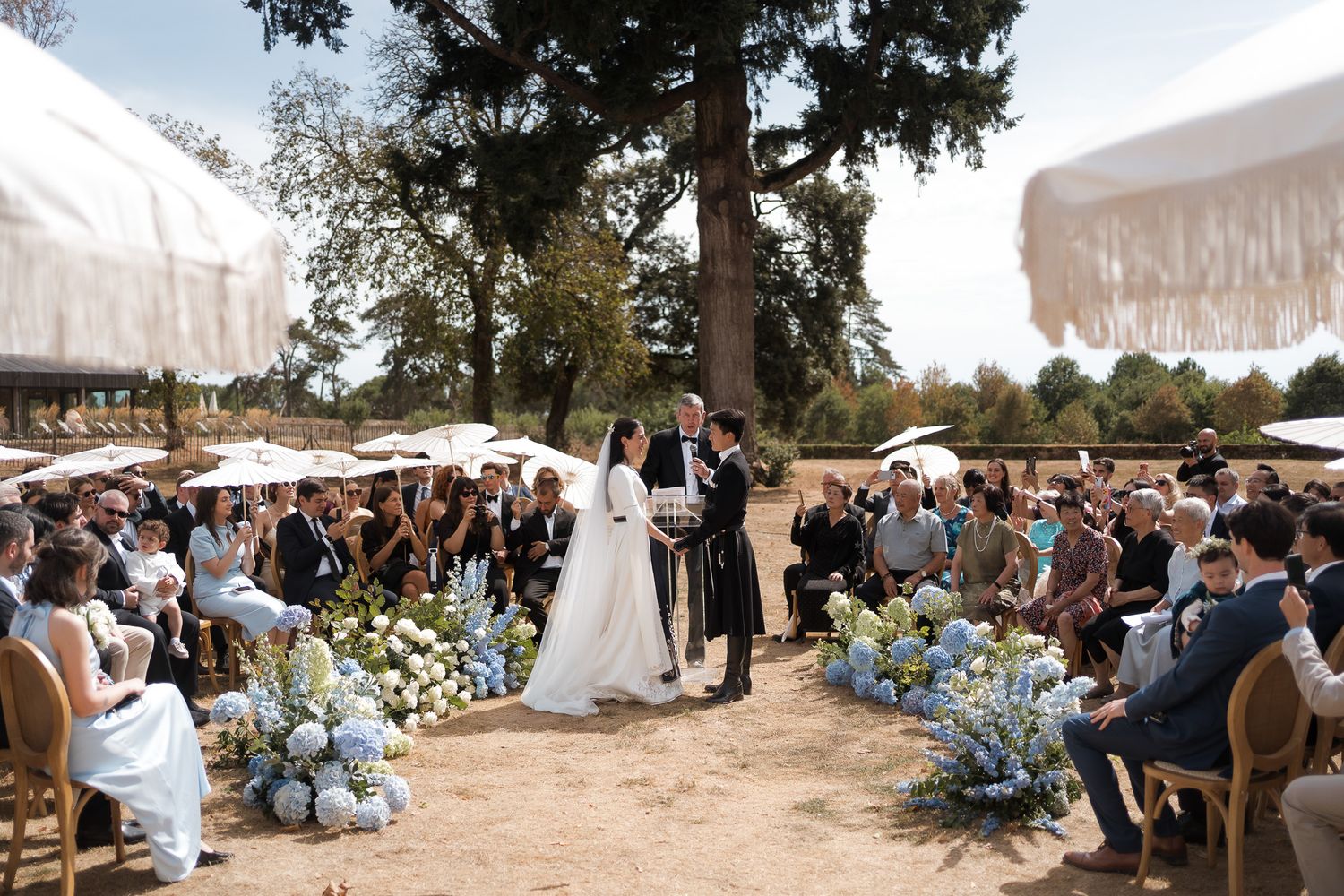 Outdoor wedding ceremony with bride and groom at altar, blue floral arrangements, white umbrellas, and seated guests among tall trees.