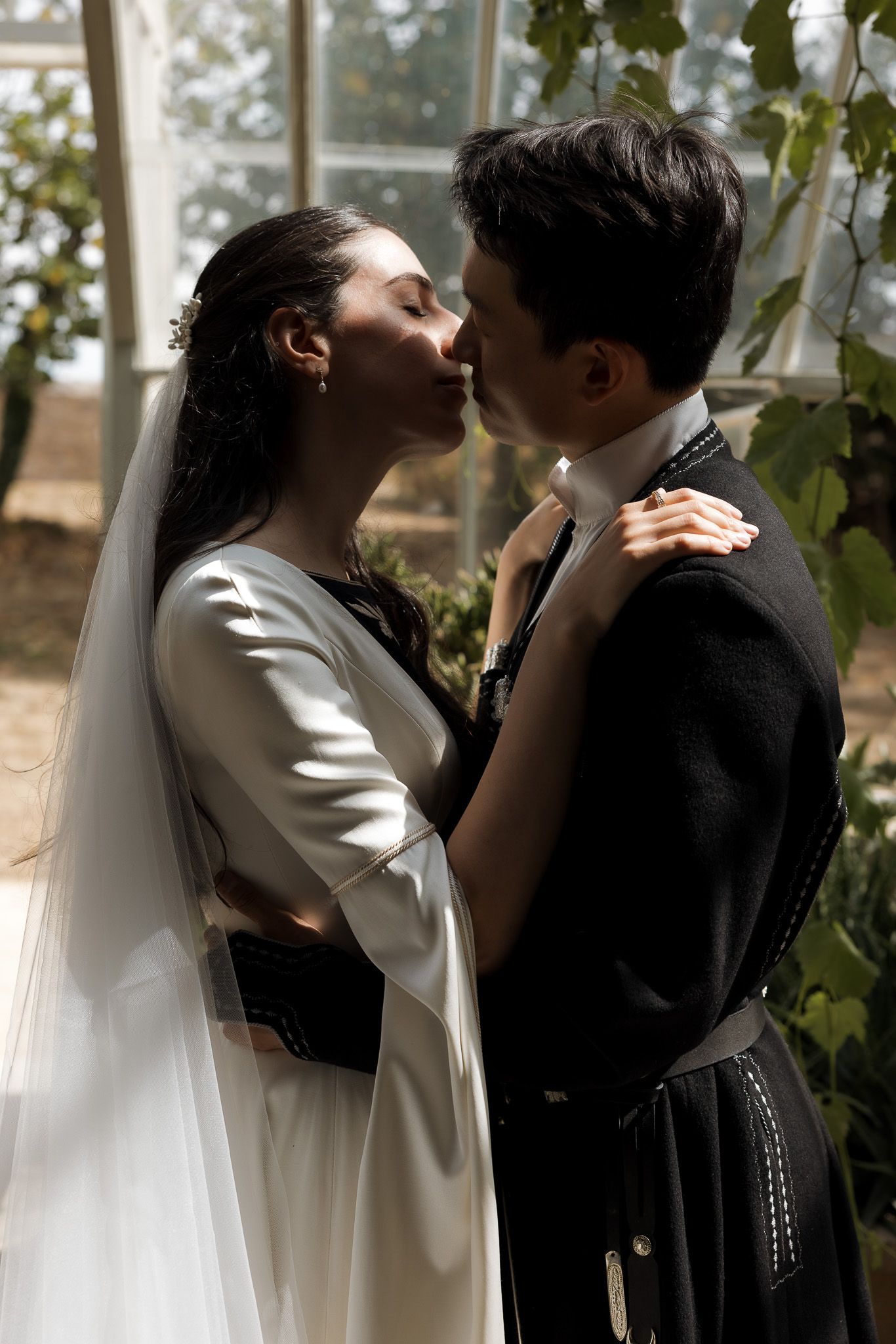 A bride and groom share a romantic kiss in a greenhouse, she wearing a white gown and veil, he in a black kilt.